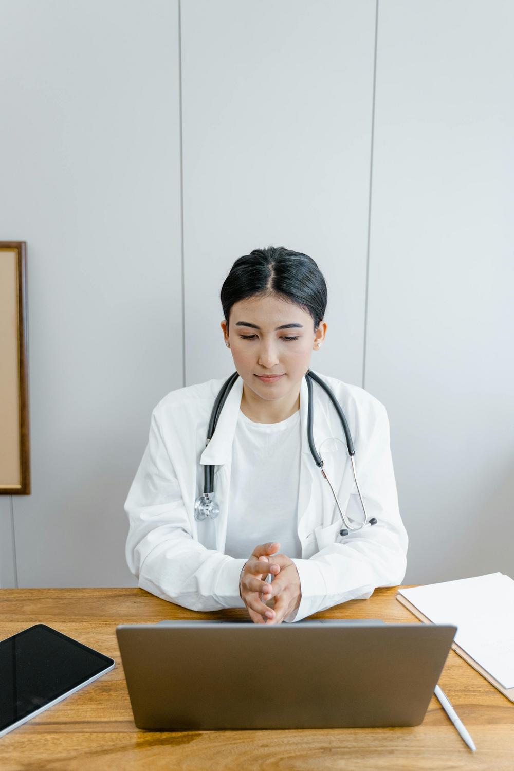 A doctor wearing a white coat and stethoscope sits at a desk, looking at a laptop with a tablet and documents nearby.