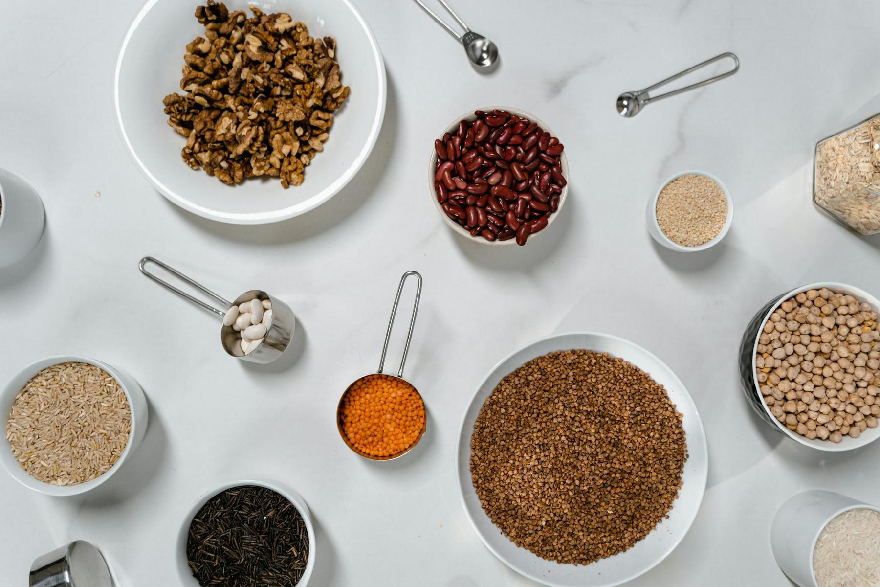 Various grains, seeds, and legumes are arranged in bowls and measuring cups on a marble surface. Items include walnuts, kidney beans, rice, black tea leaves, chickpeas, and quinoa.