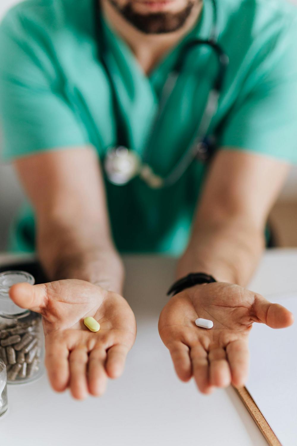 A person in medical scrubs with a stethoscope holds a yellow pill in one hand and a white pill in the other hand at a desk with a pill bottle.