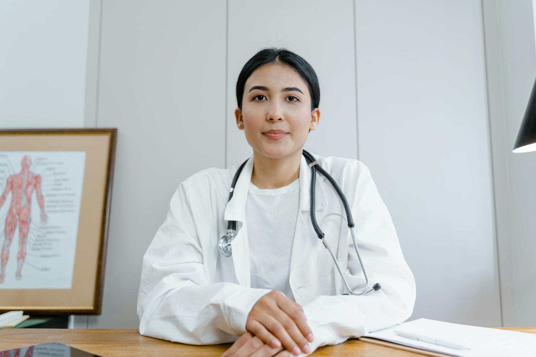 A doctor in a white coat with a stethoscope sits at a desk in a medical office, with an anatomical chart in the background.