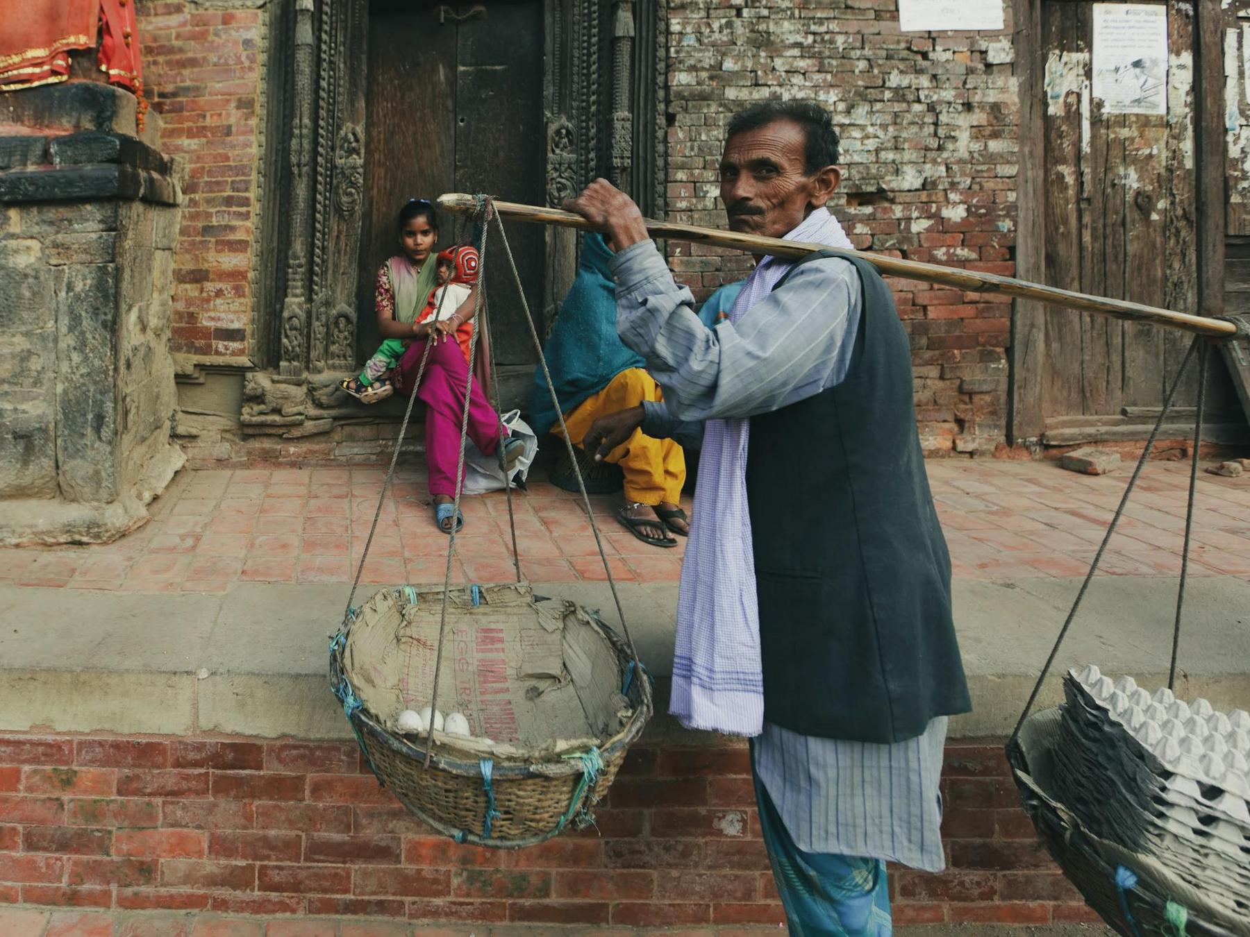 A man carries baskets with eggs using a wooden pole on his shoulder, while two women sit on a brick platform in front of an old building.