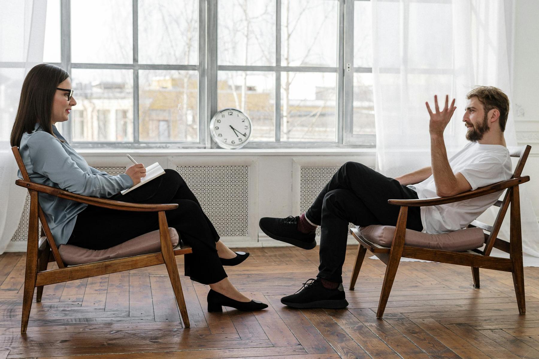 A woman with a notepad listens to a man speaking with a raised hand; they sit in chairs facing each other in a bright room with a clock on the windowsill.
