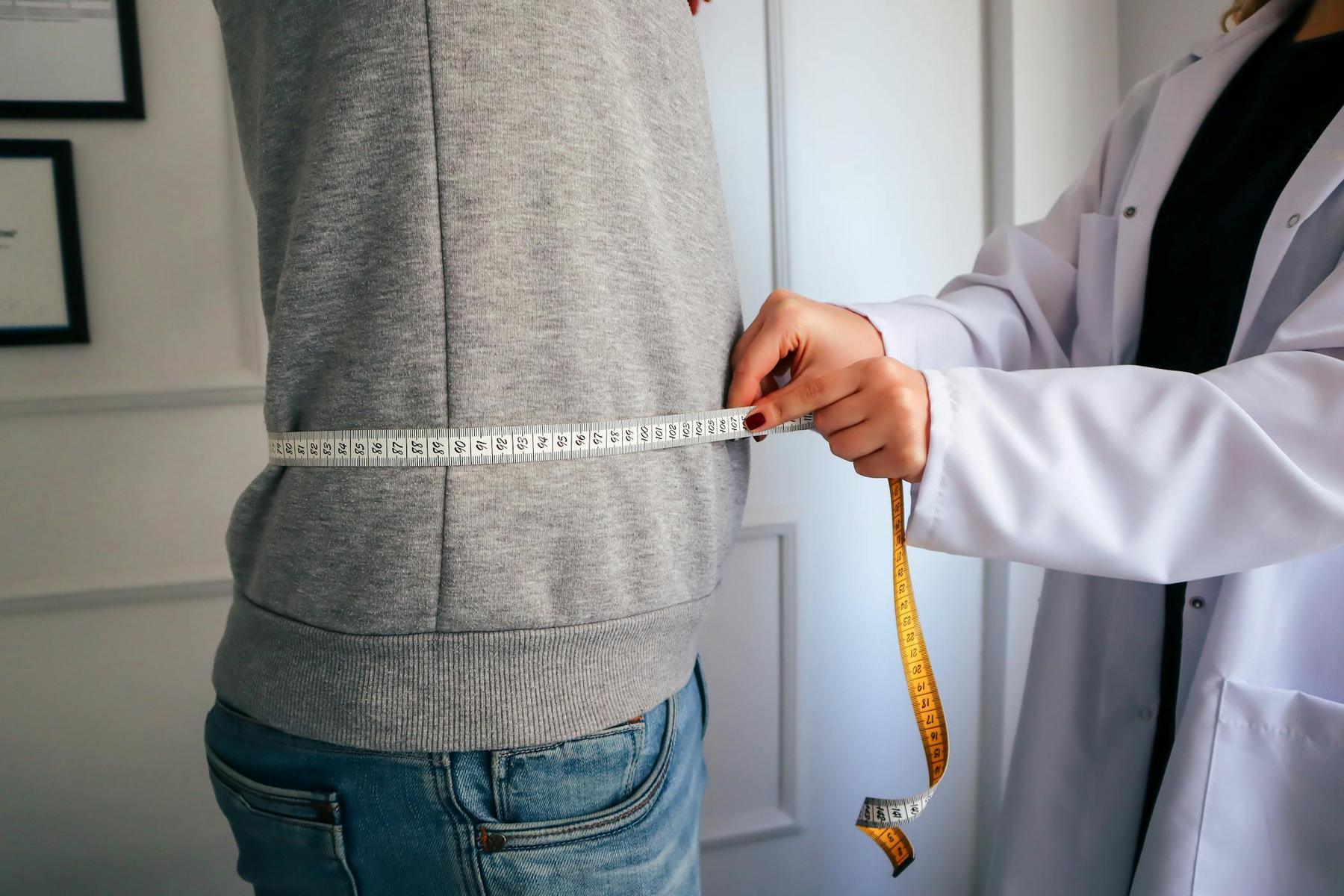 A person in a gray sweater is having their waist measured with a tape measure by someone in a white coat.