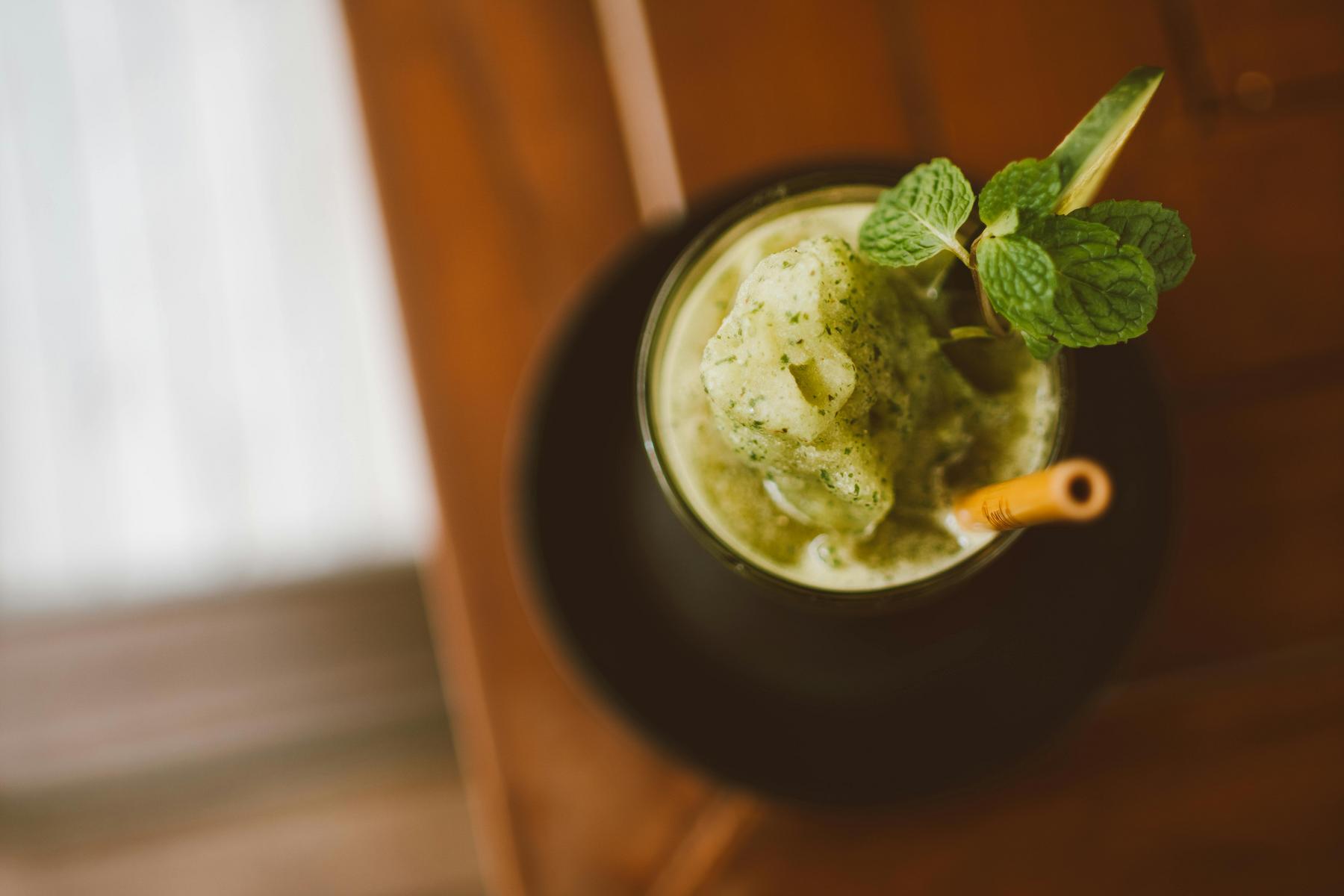Overhead view of a green smoothie with a straw in a glass, garnished with mint leaves, placed on a wooden table.