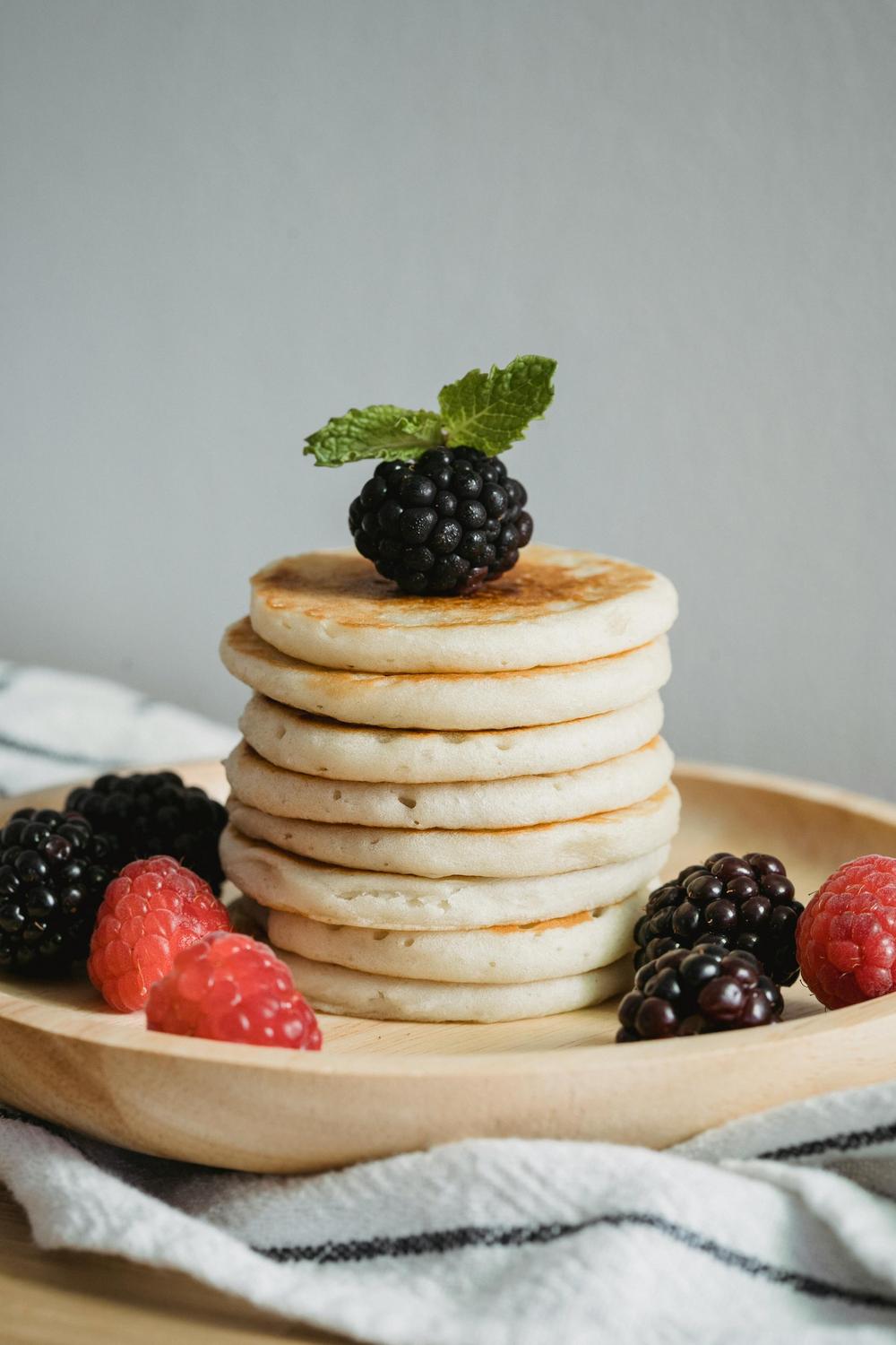 A stack of six pancakes on a wooden plate, topped with a blackberry and mint leaf, surrounded by blackberries and raspberries.