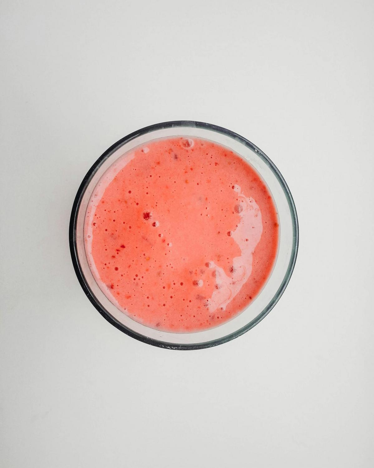 A glass filled with a pink, frothy liquid, likely a fruit smoothie, photographed from above on a white background.