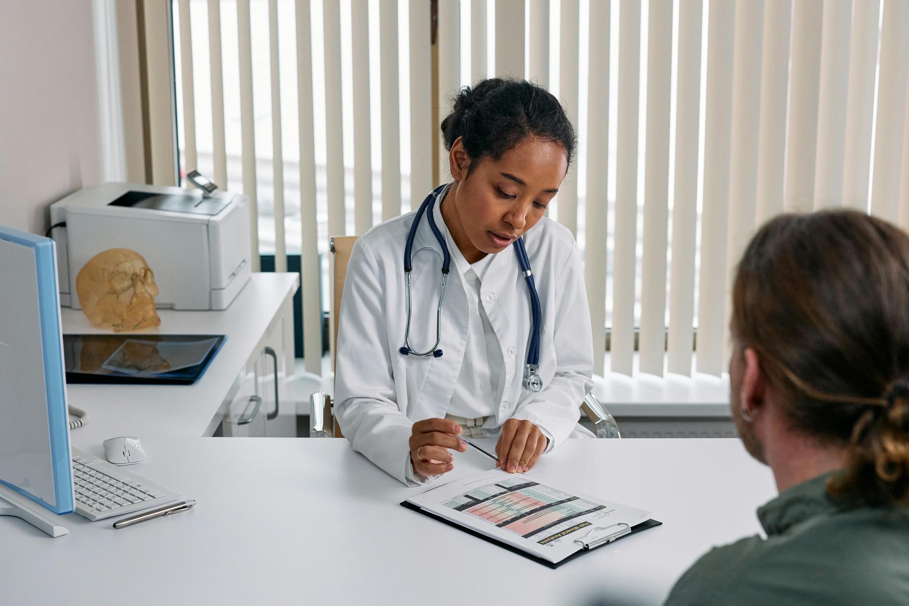 A doctor in a white coat reviews a medical chart with a patient at a desk in a well-lit office with vertical blinds.