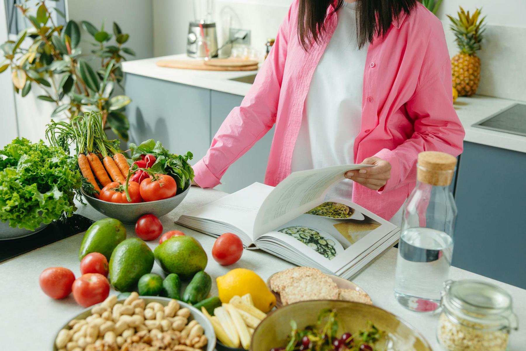 Person in a pink shirt stands in a kitchen, looking at an open cookbook, surrounded by fresh vegetables, fruits, nuts, bread, and a bottle of water on the counter.