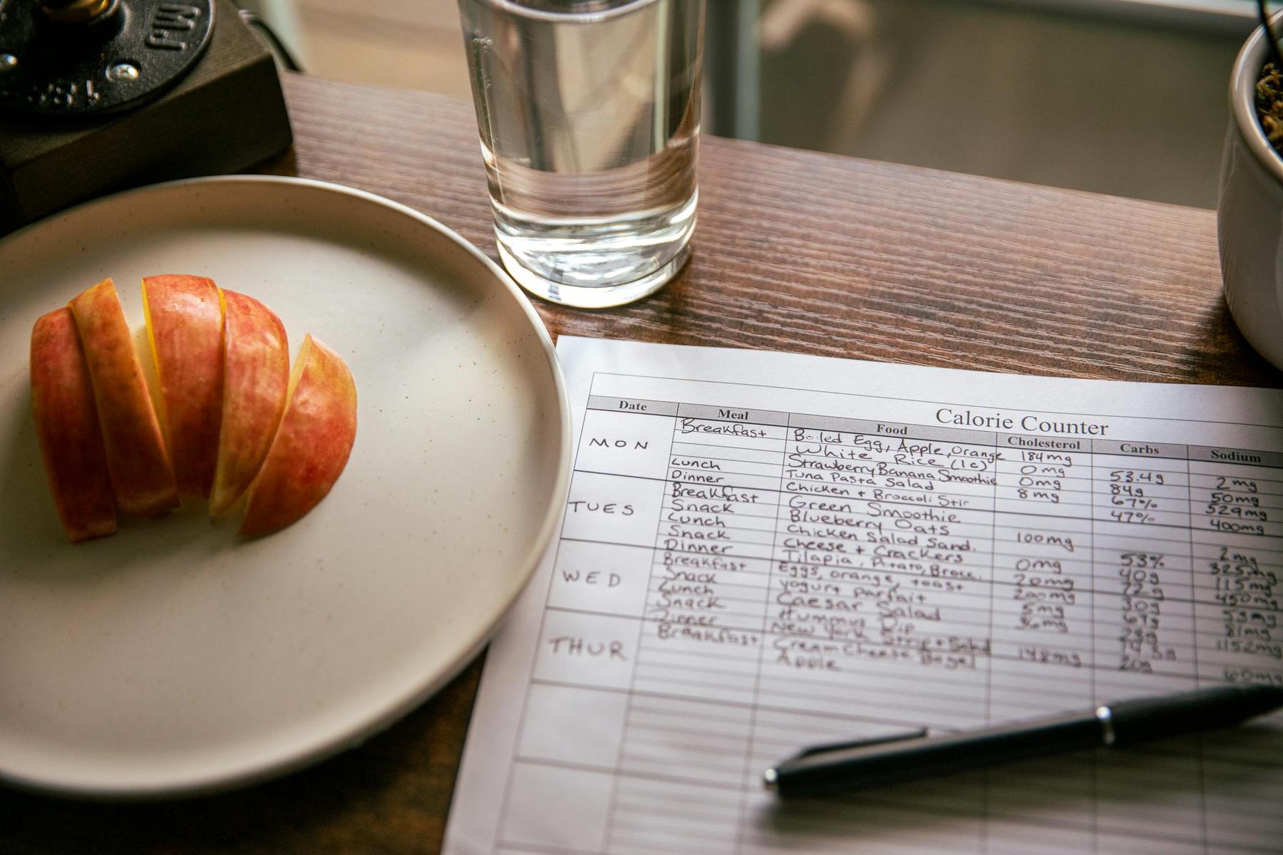 A plate with sliced apple, a glass of water, and a pen lie next to a calorie counter meal planner on a wooden table.