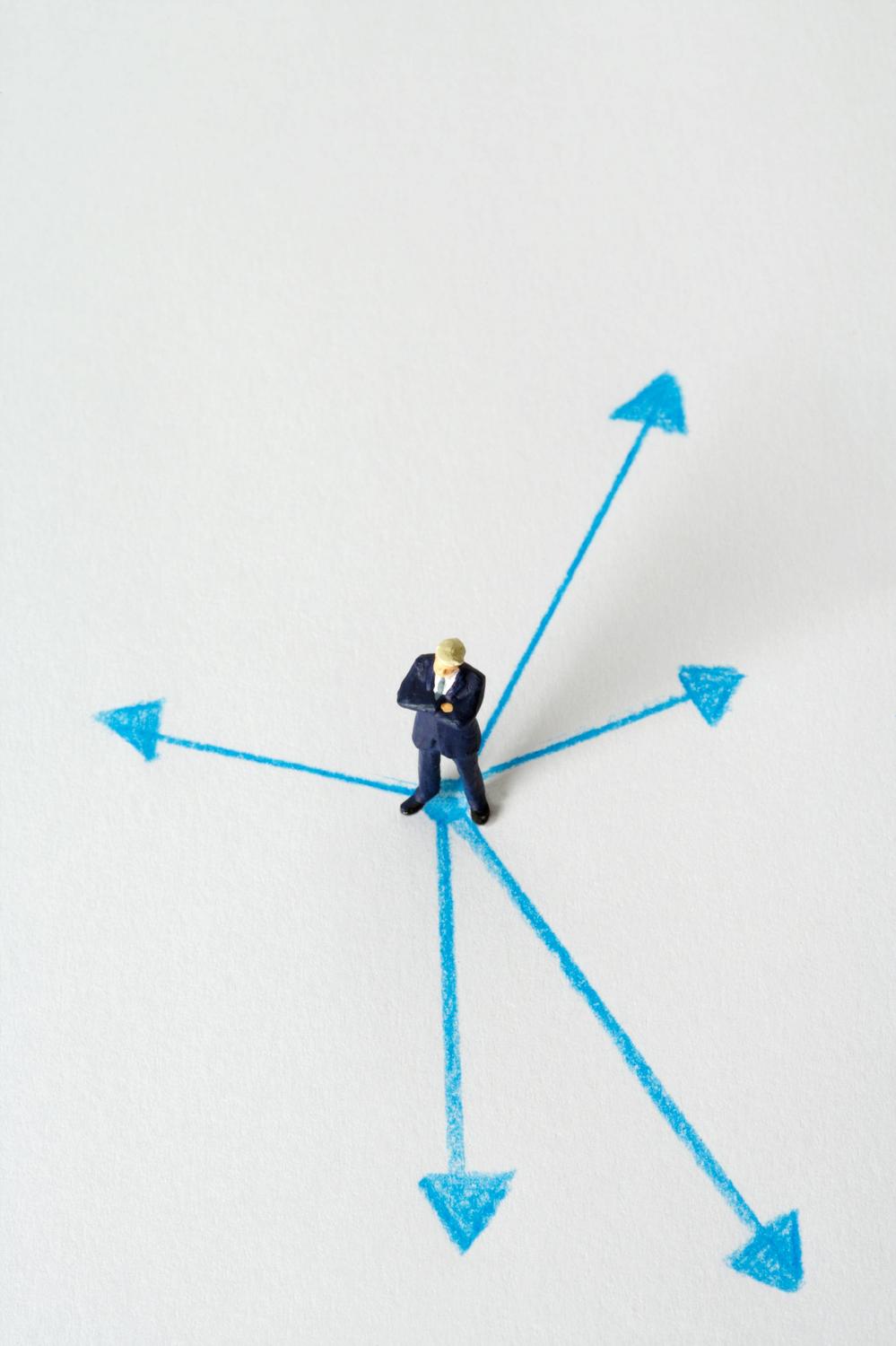 Miniature figure of a person in a suit stands at the intersection of several blue arrows pointing in different directions on a white background.