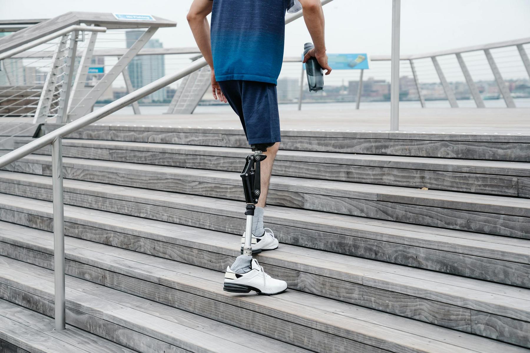 A person with a prosthetic leg, dressed in athletic wear, walks up outdoor wooden stairs while holding a water bottle.