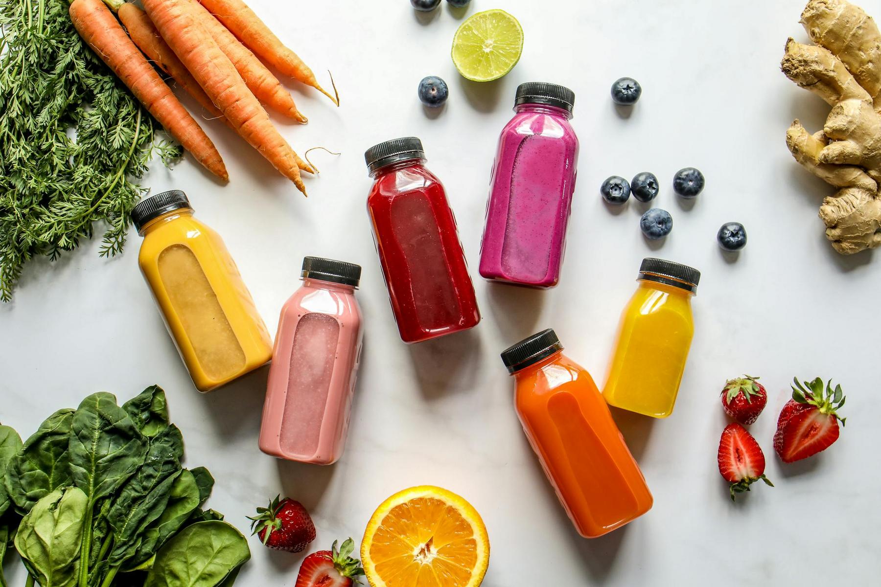 Six bottles of assorted colorful juices are arranged on a white surface, surrounded by fresh fruits, vegetables, ginger, carrots, spinach, blueberries, strawberries, and citrus.