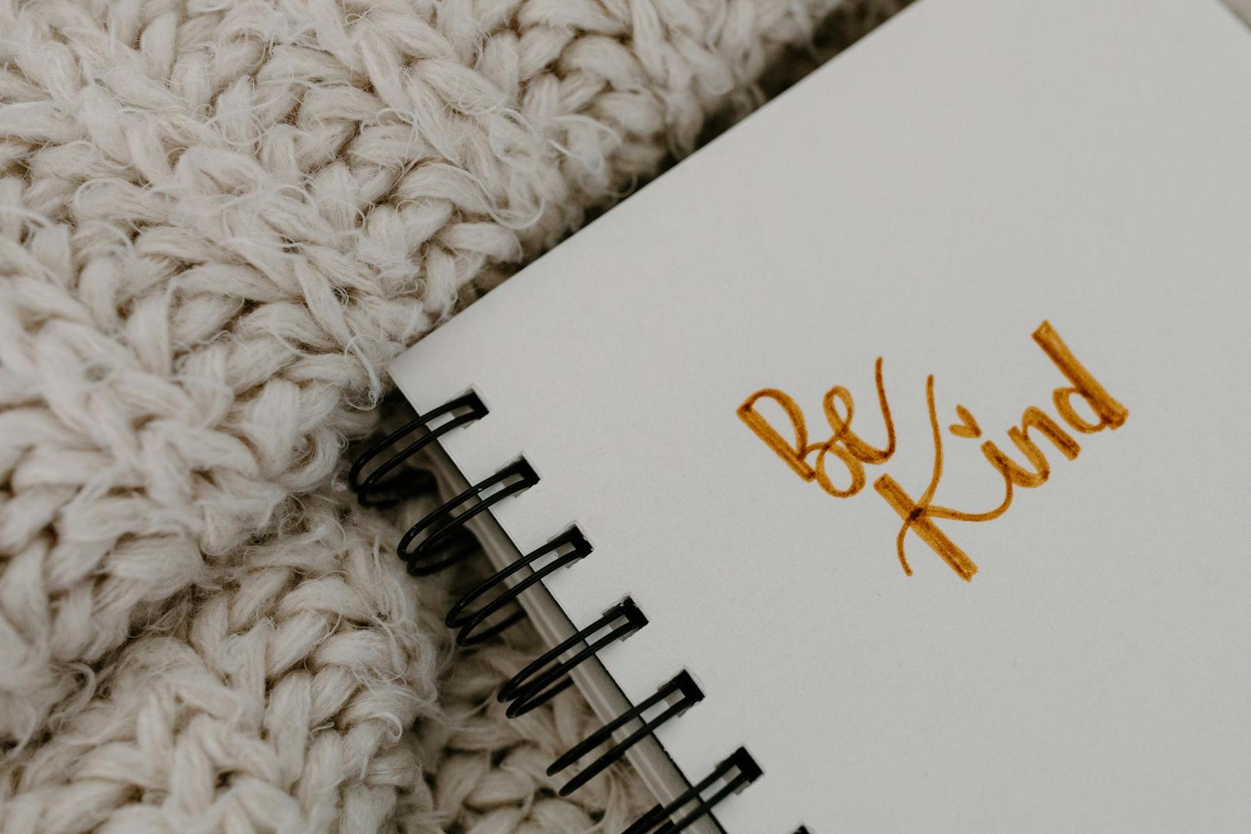 A close-up of a spiral notebook with “Be Kind” written in yellow on the cover, next to a chunky knitted blanket.