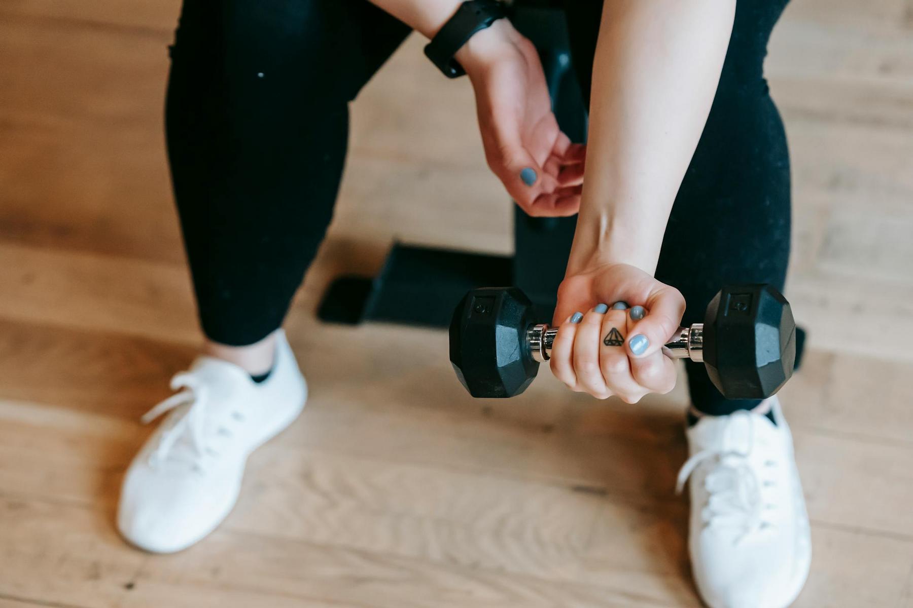 Person wearing black leggings and white sneakers sitting on a bench, holding a single dumbbell in one hand over a wooden floor.