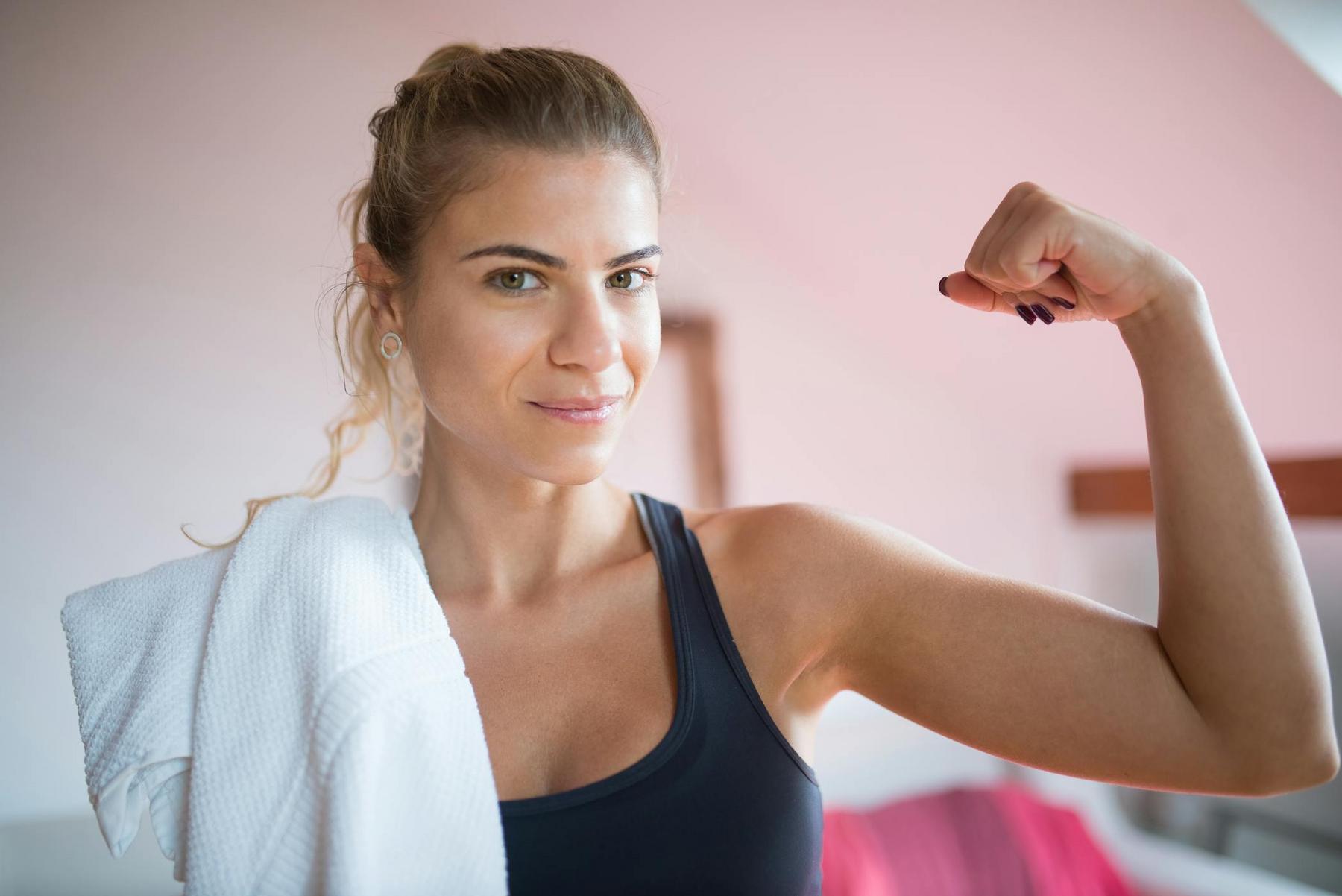 Woman in athletic wear flexes her arm and holds a white towel over her shoulder, looking confidently at the camera.