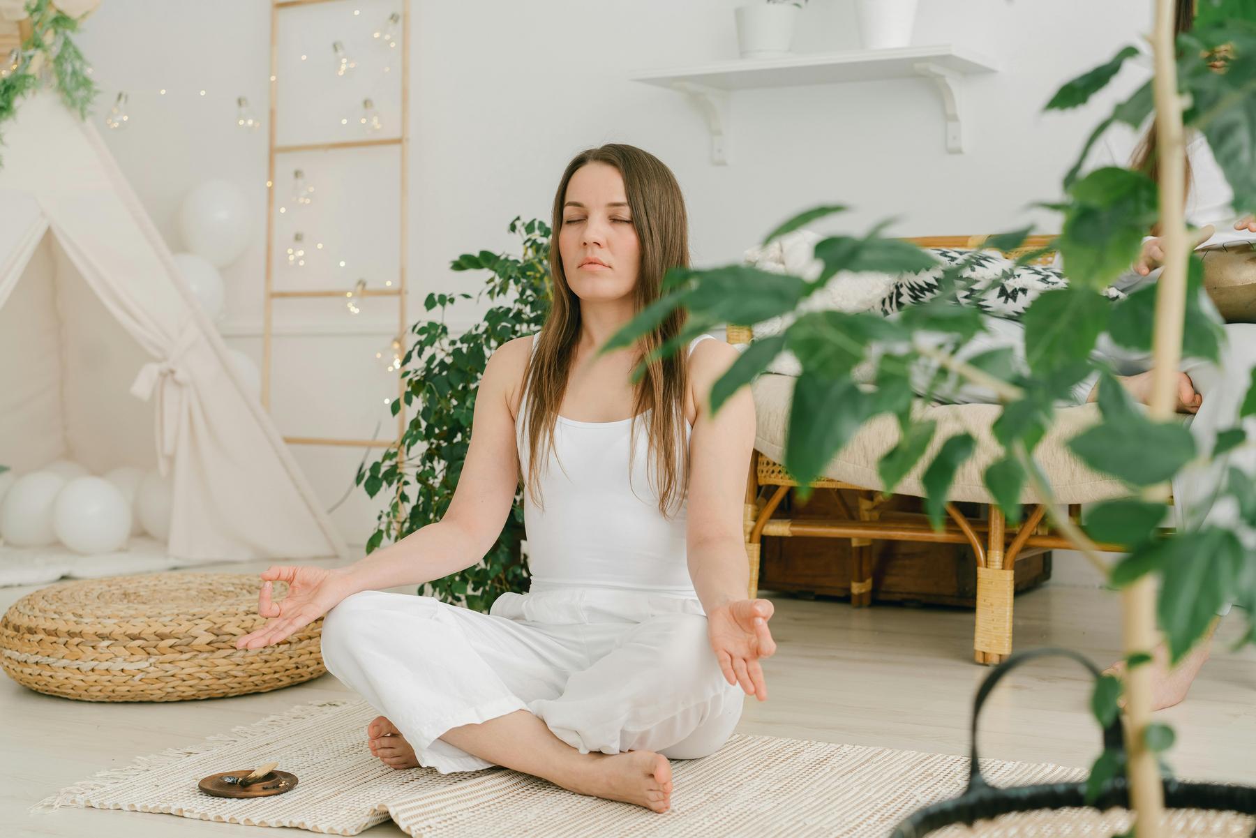 A woman sitting cross-legged on a rug in a serene, plant-filled room, meditating with closed eyes and relaxed hands.