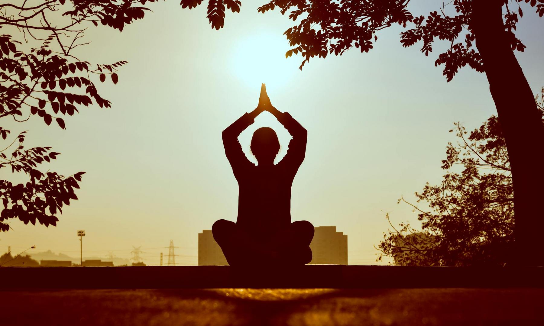 A person sits cross-legged outdoors at sunrise or sunset, holding hands together above their head in a yoga pose, with trees and buildings in the background.