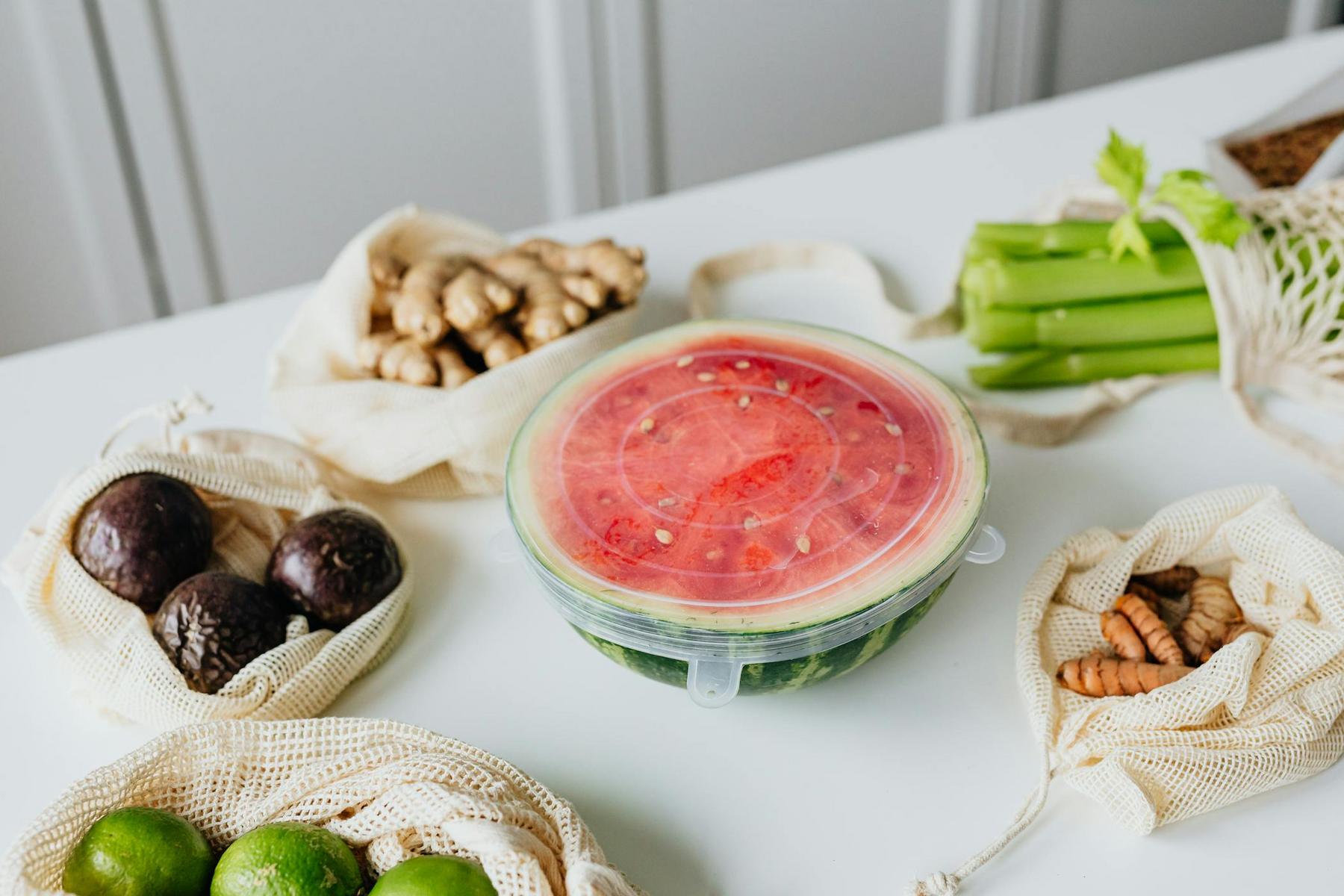 A round glass container with watermelon slices, covered by a silicone lid, surrounded by bags of ginger, avocados, celery, turmeric, and limes on a white surface.