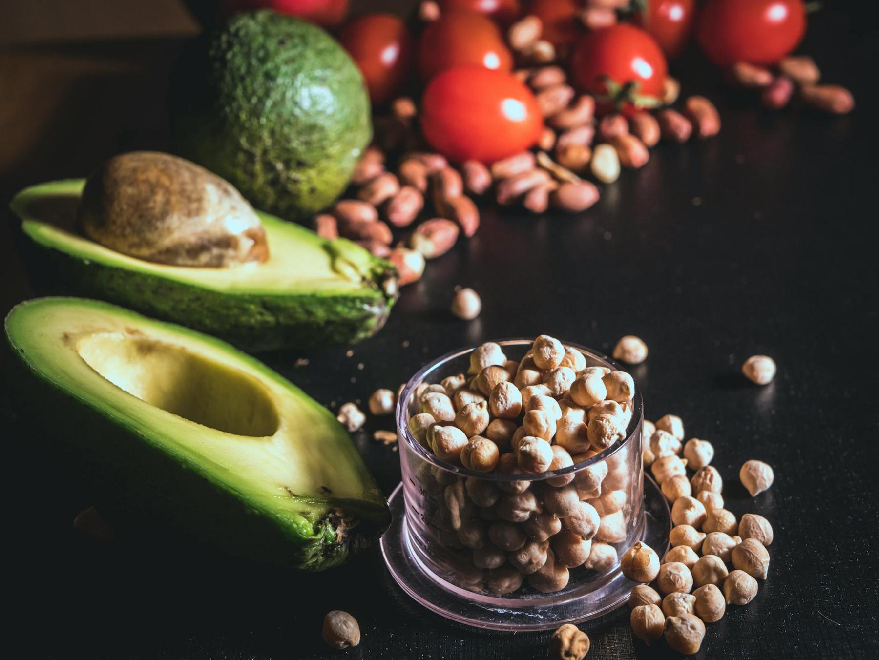 A halved avocado, cherry tomatoes, peanuts, and chickpeas in a small cup are arranged on a dark surface.