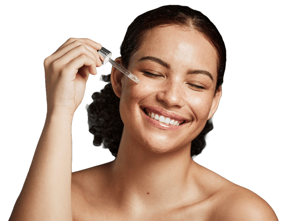 A woman with closed eyes and a smile applies a drop of serum to her cheek using a dropper against a plain white background.