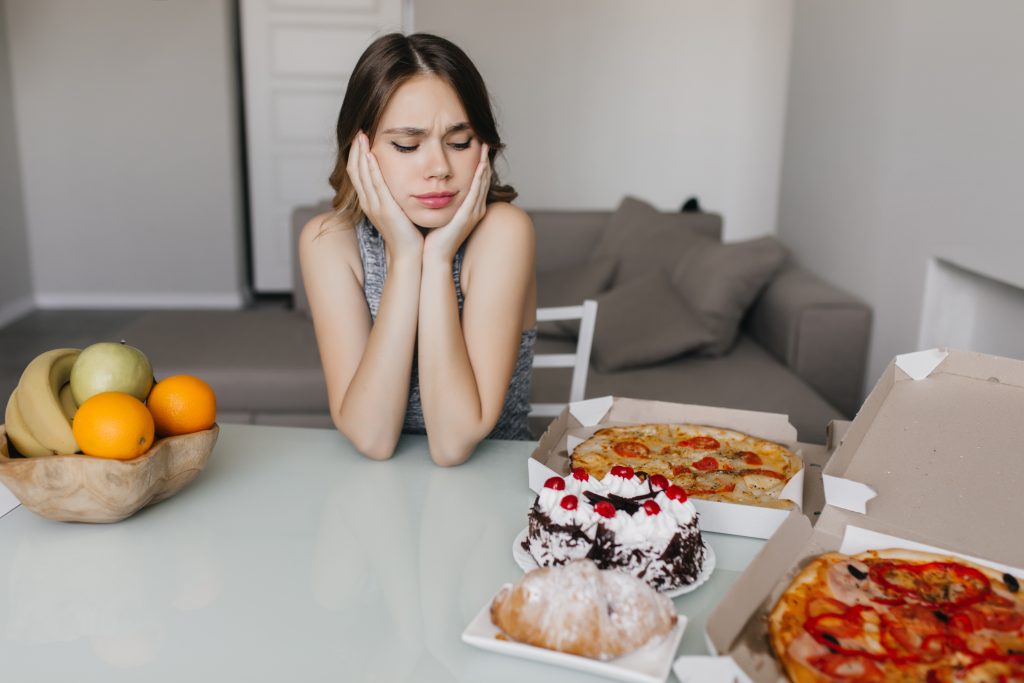 A person sits at a table with a thoughtful expression, surrounded by pizzas, pastries, and a bowl of fruit, pondering the connection between indulgence and weight loss.