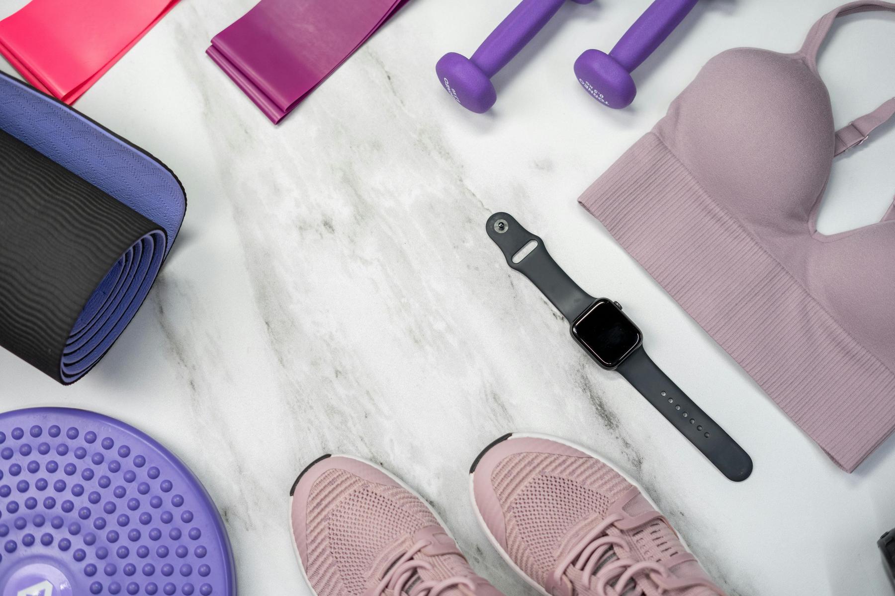 Workout gear on a marble surface, including pink sneakers, a sports bra, resistance bands, dumbbells, an exercise mat, a balance disc, and a smartwatch.