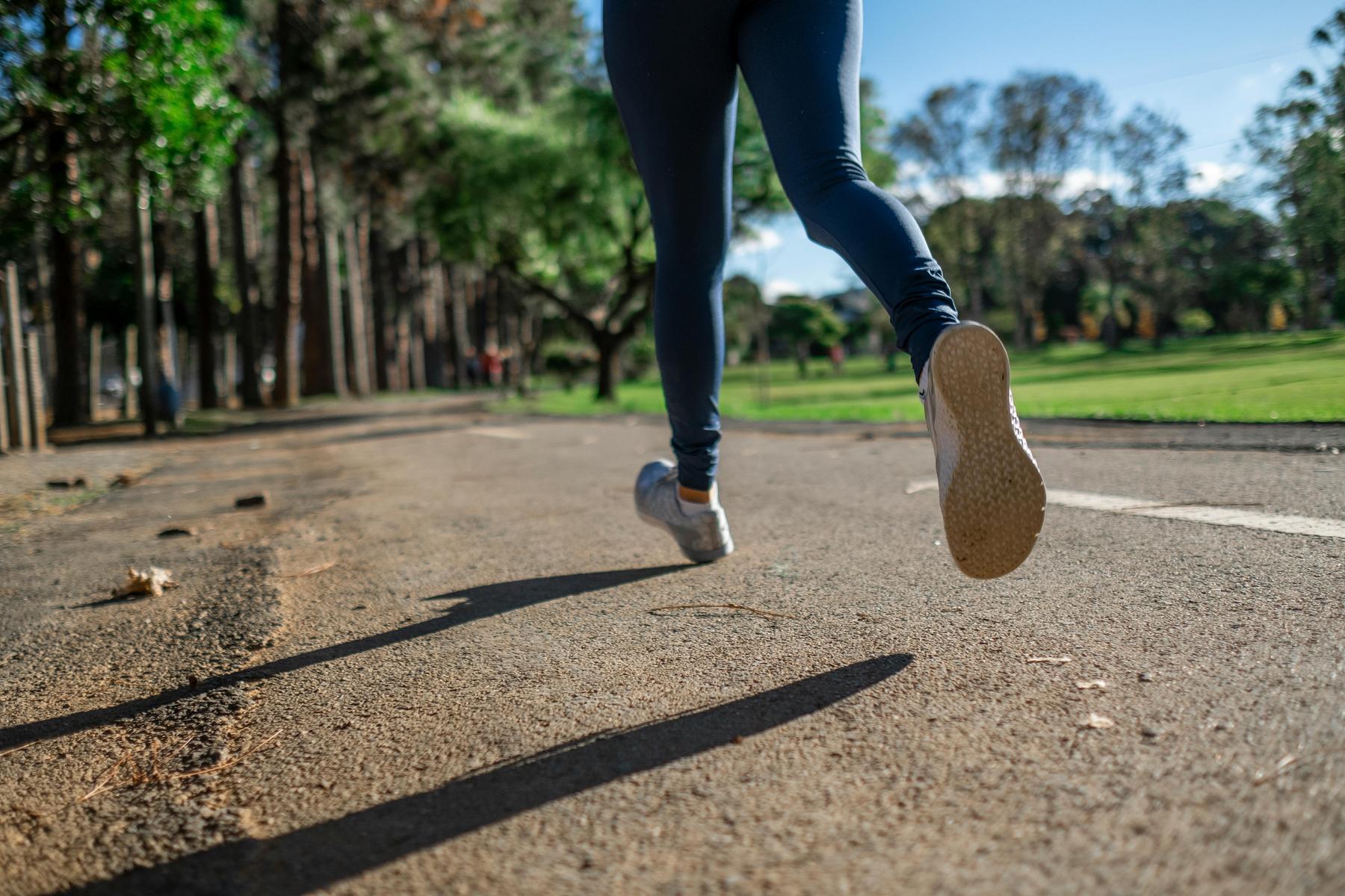 Person in athletic wear jogging on a paved path in a park surrounded by trees and greenery on a sunny day.