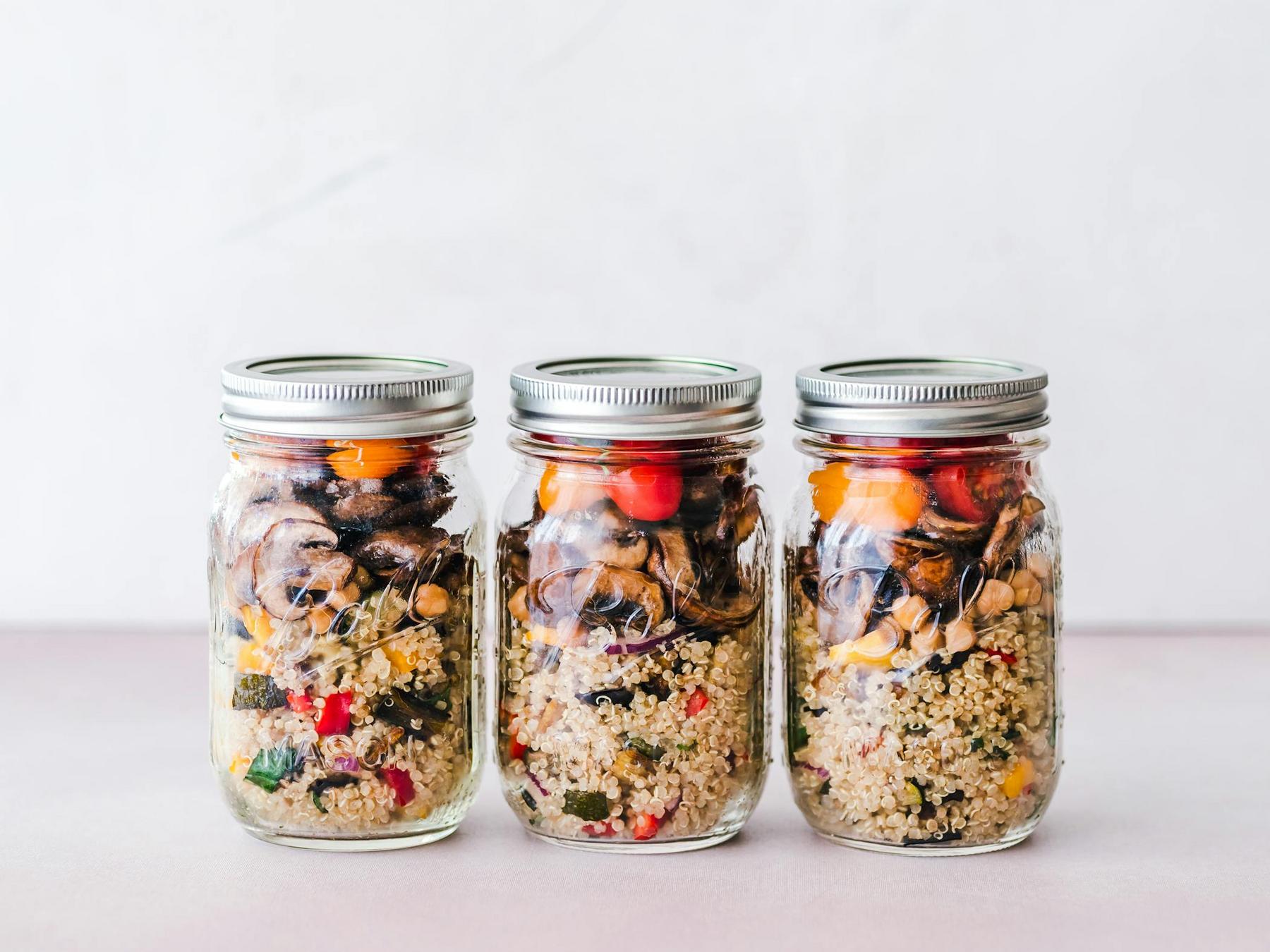 Three glass jars filled with layered quinoa, roasted vegetables, and chickpeas, each sealed with a metal lid, are arranged side by side on a light surface.