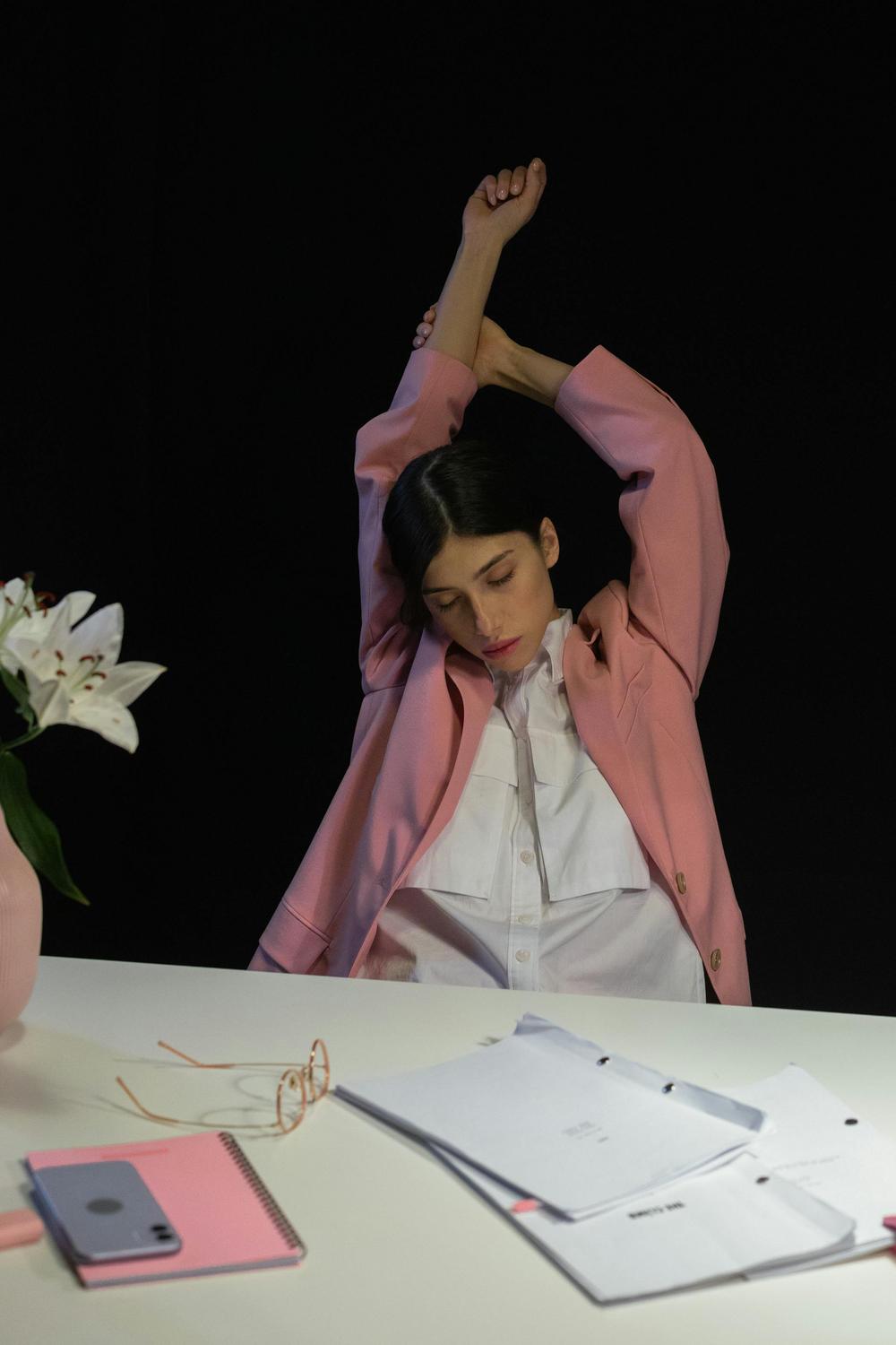 A woman in a pink blazer sits at a desk, stretching with arms raised. Open notebooks, a pen, and a vase with white lilies are on the desk.