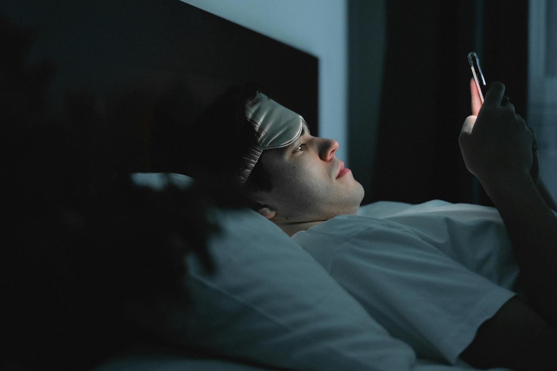 A person lying in bed at night looks at a smartphone screen while wearing a sleep mask on their forehead.