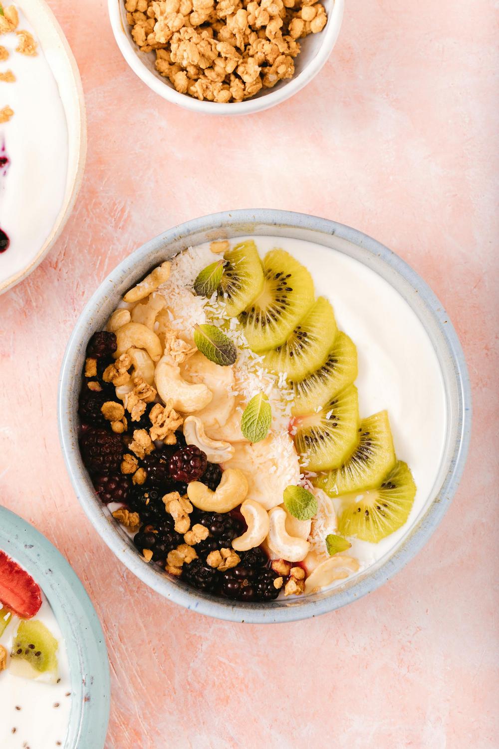 A bowl of yogurt topped with sliced kiwi, blackberries, cashews, granola, coconut flakes, and mint leaves, on a light pink surface.