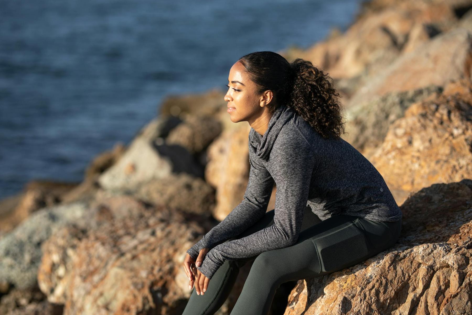 Woman in athletic wear sitting on rocks by the water, looking into the distance.