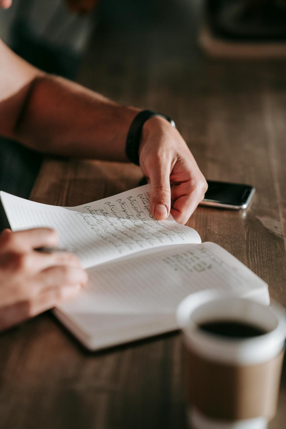 A person writes in a notebook on a wooden table, with a smartphone and a cup of coffee nearby.