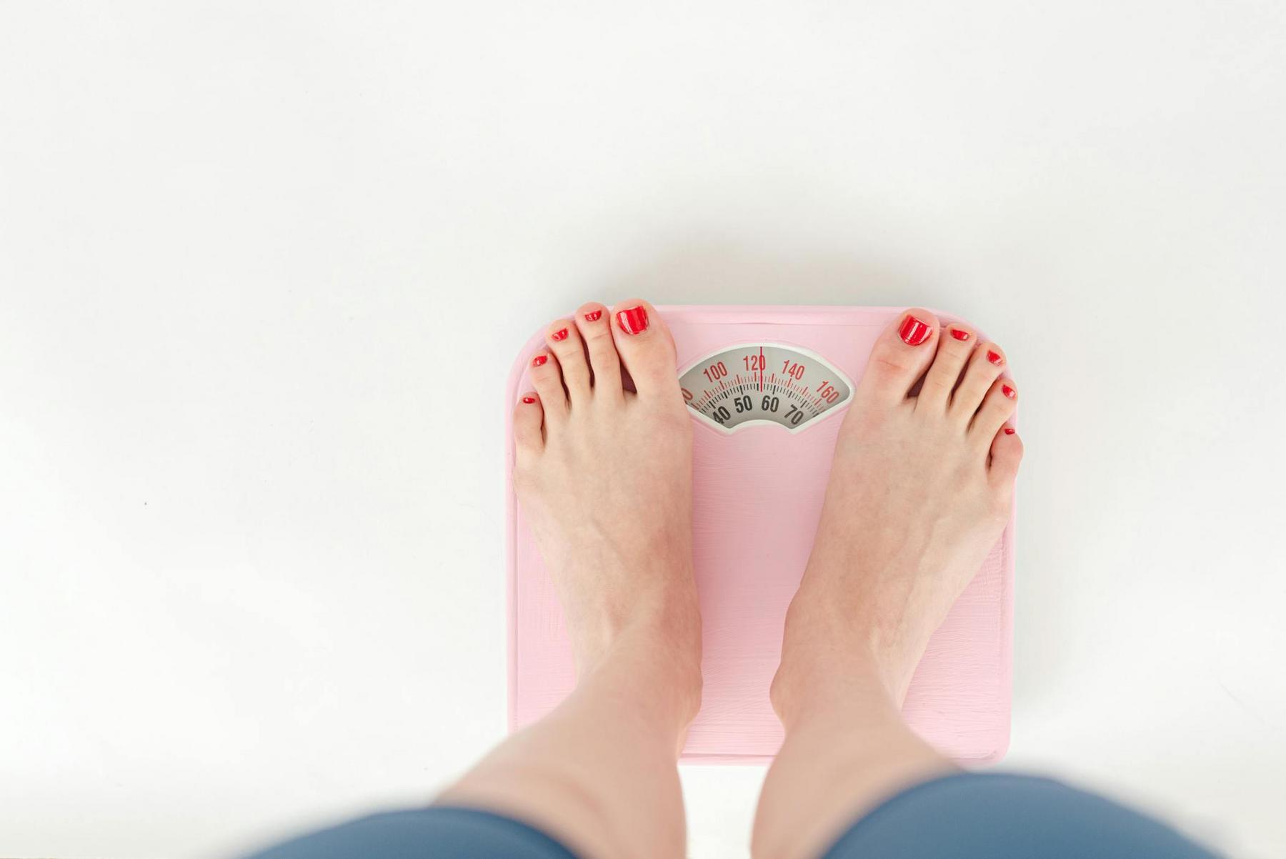 Person standing on a pink analog scale showing weight, with feet visible, toenails painted red, against a white background.