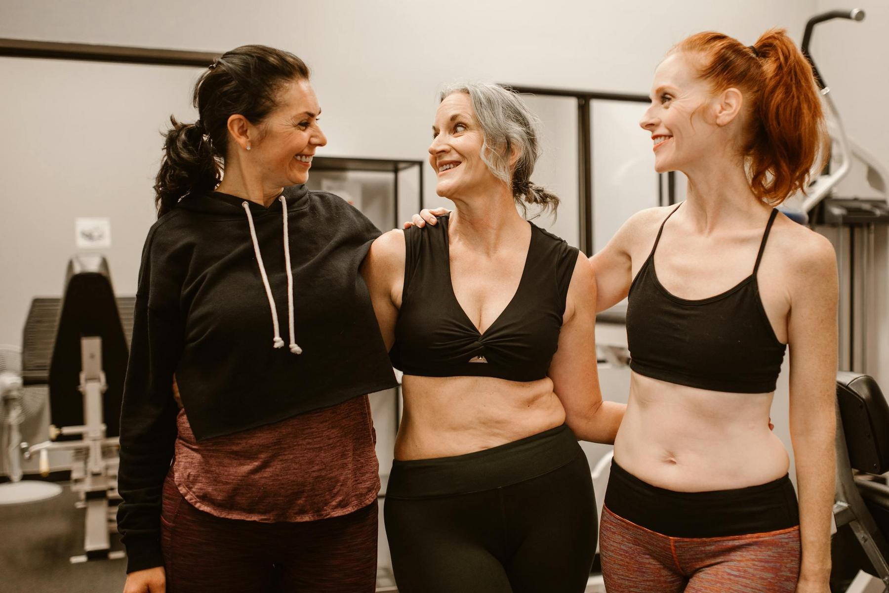 Three women in activewear stand in a gym, smiling and linking arms, with gym equipment visible in the background.