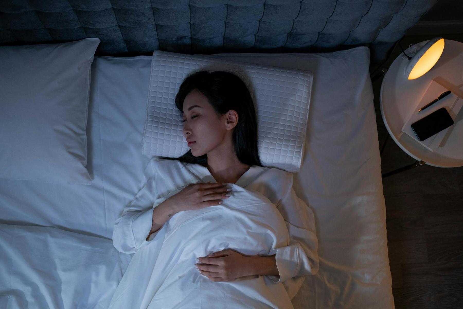 A woman lies on her back in bed, eyes closed, with one hand on her chest. A lit lamp and a phone are on the bedside table next to her.