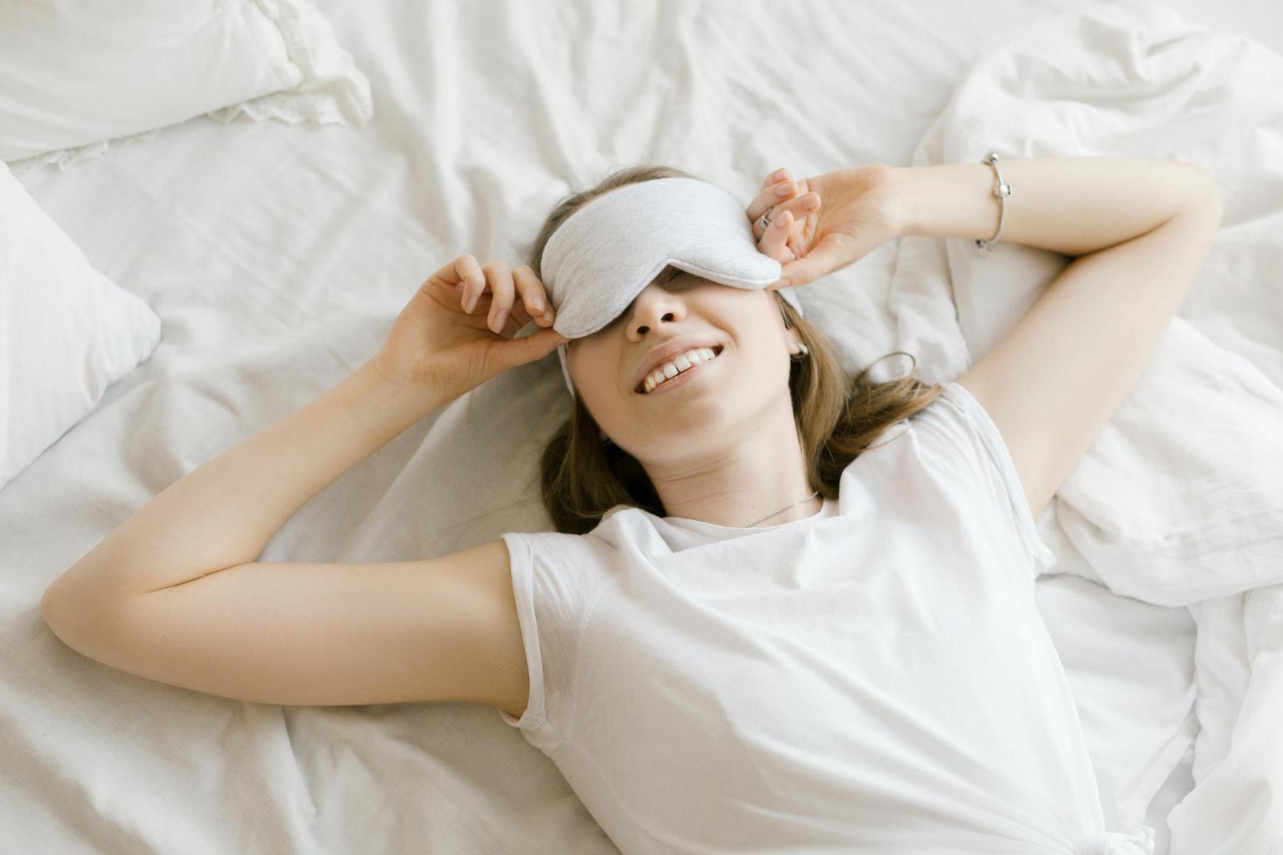 Person lying on a bed wearing a sleep mask, smiling, with arms raised and resting on the pillow.