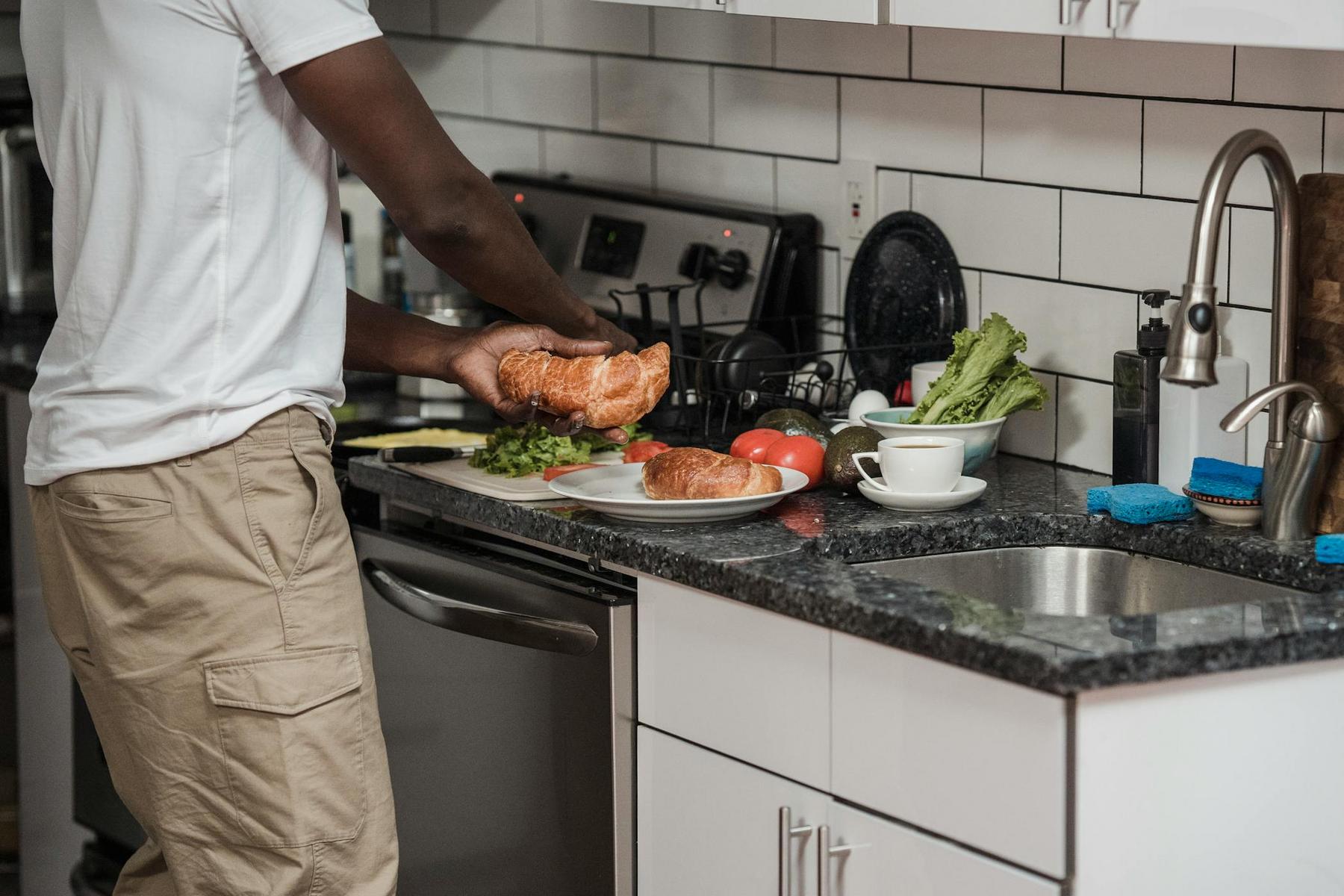 A person in a white shirt and khaki pants prepares food on a kitchen counter with lettuce, tomatoes, and bread next to a cup and plate.