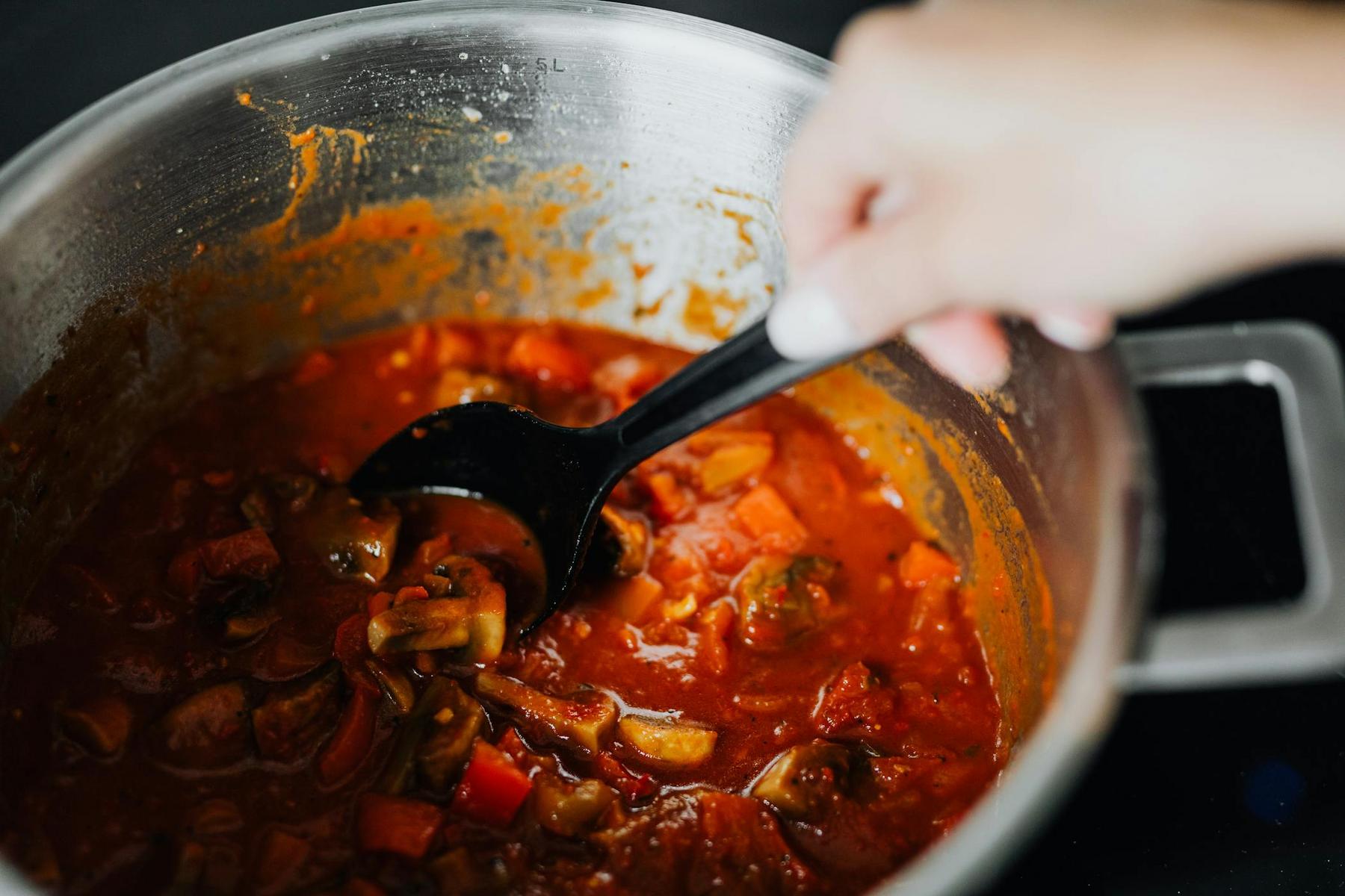 A hand stirring a pot of tomato-based stew with vegetables using a black ladle.