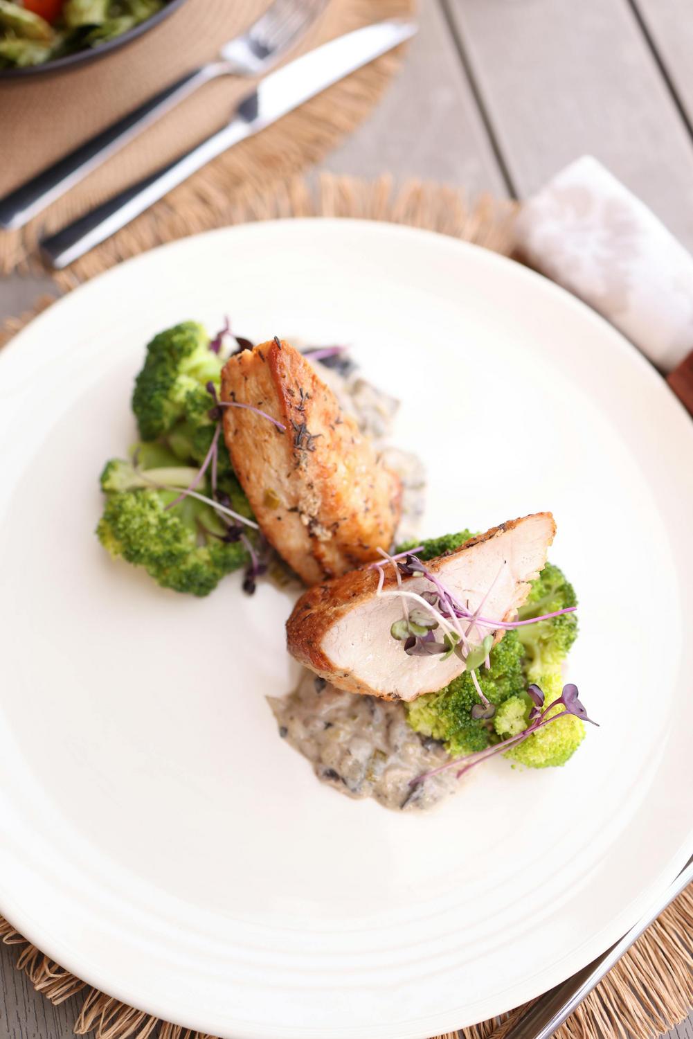 Sliced roasted chicken breast served with broccoli and a creamy mushroom sauce, garnished with microgreens on a white plate.