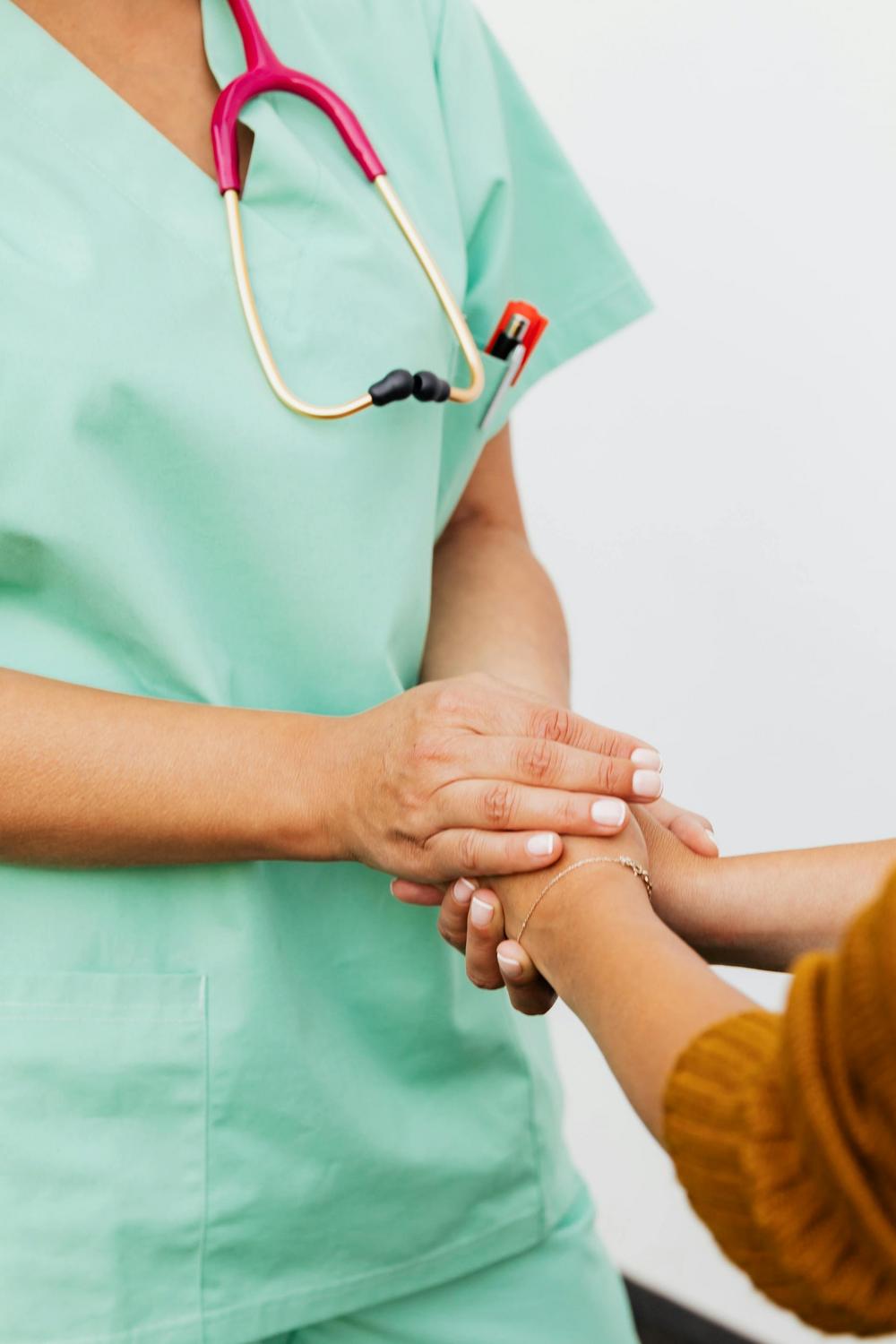 A healthcare worker in green scrubs with a stethoscope holds the hand of a patient, offering support.