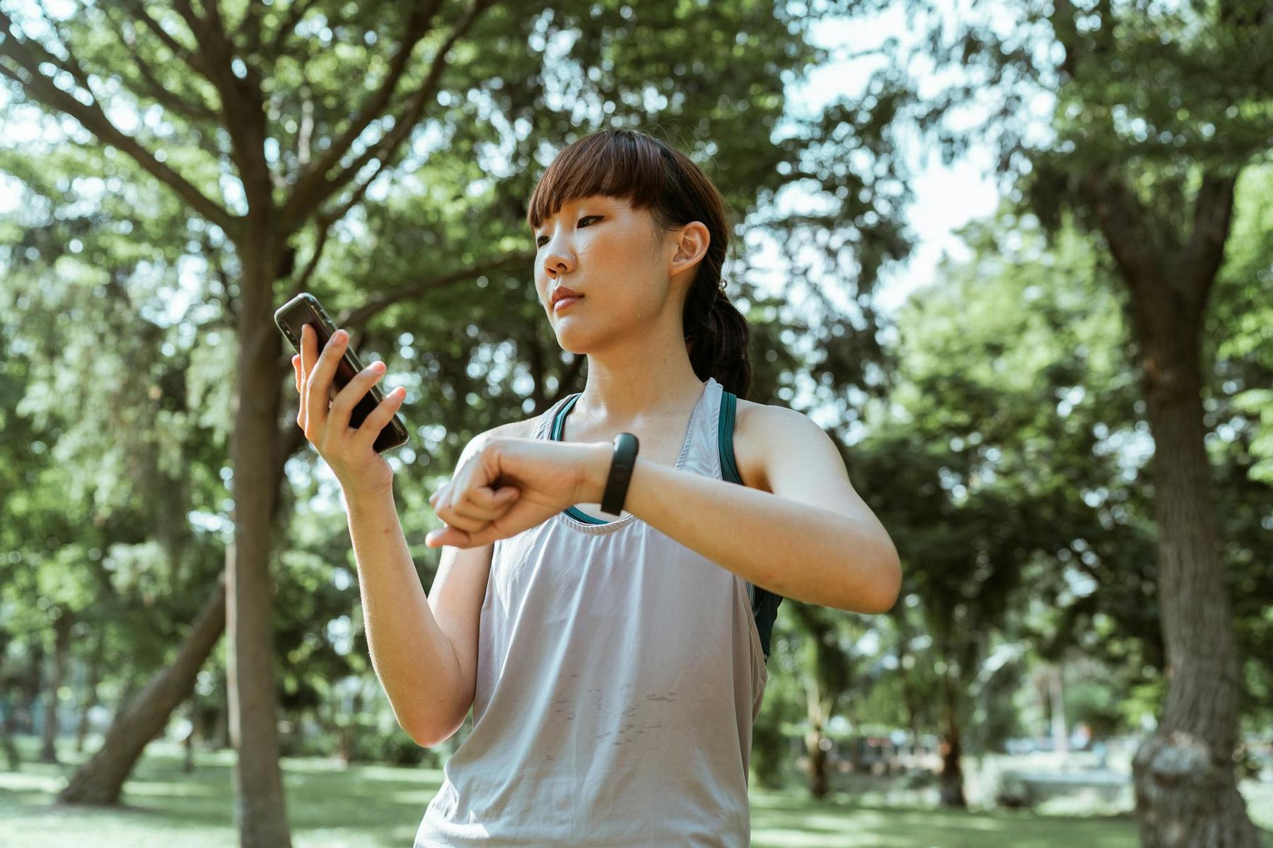 Person in athletic attire checks smartphone and wrist device in a park with trees in the background.