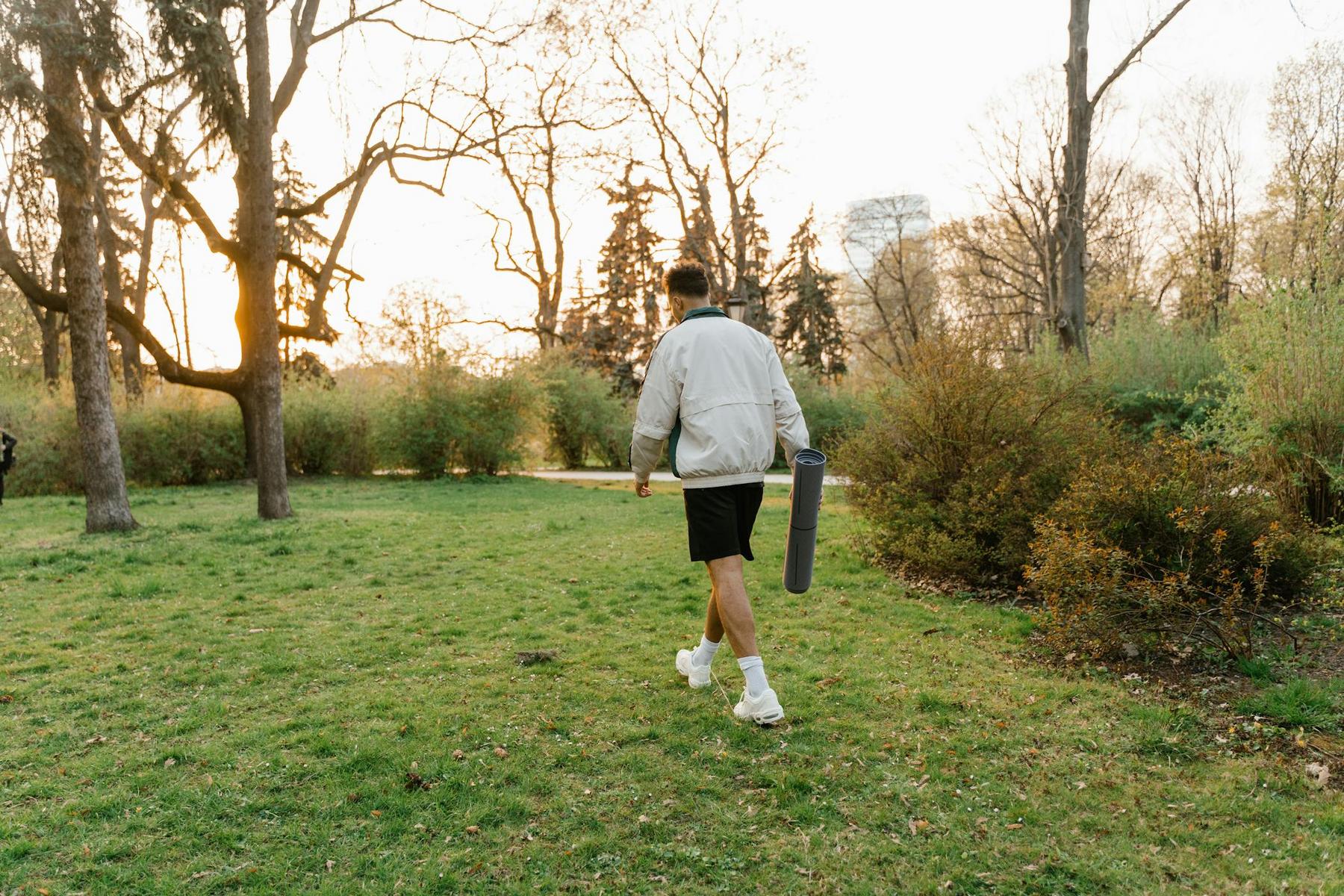 A person in athletic wear walks across a grassy park carrying a yoga mat, with trees and bushes in the background at sunset.