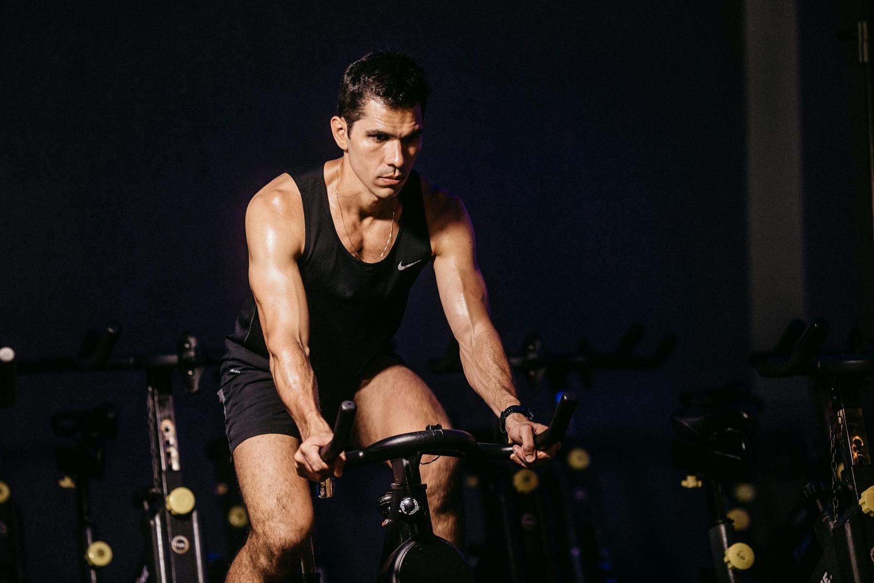 A man wearing a black tank top and shorts is intensely riding a stationary exercise bike in a dimly lit gym.