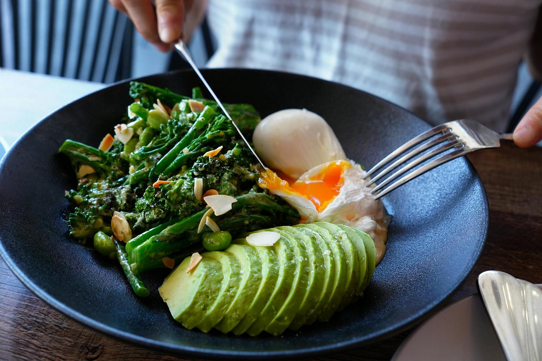 A person cuts into a poached egg with a knife and fork, revealing a runny yolk. The dish includes broccoli, sliced avocado, and almond slivers on a black plate.