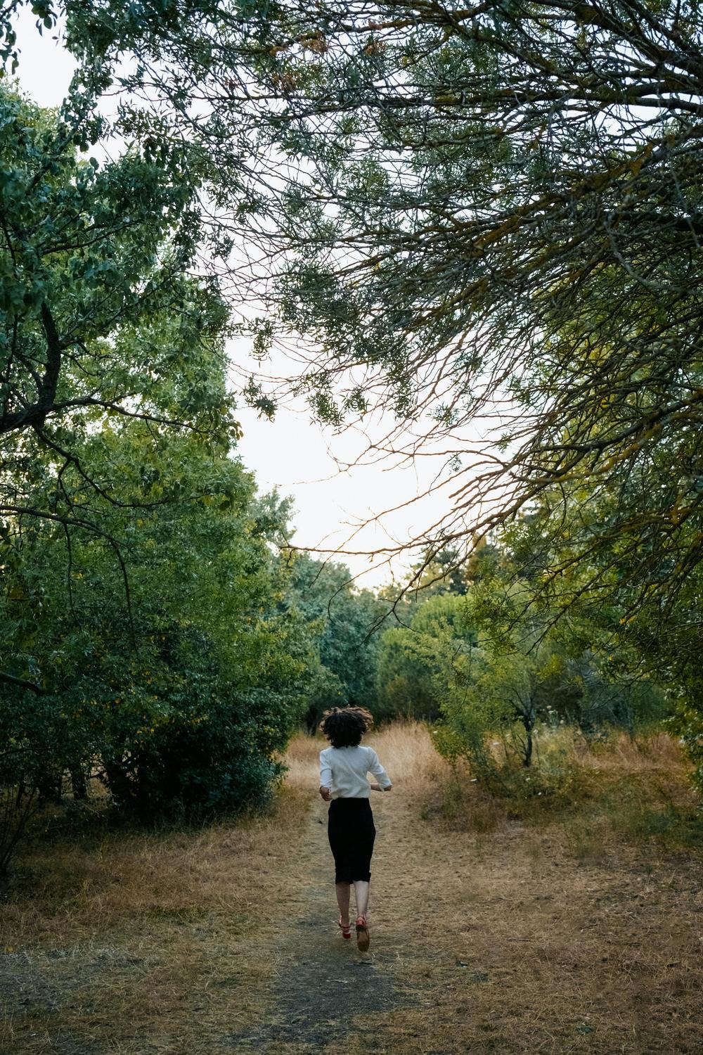A person with curly hair walks alone on a dirt path through a forested area with green trees and dry grass.