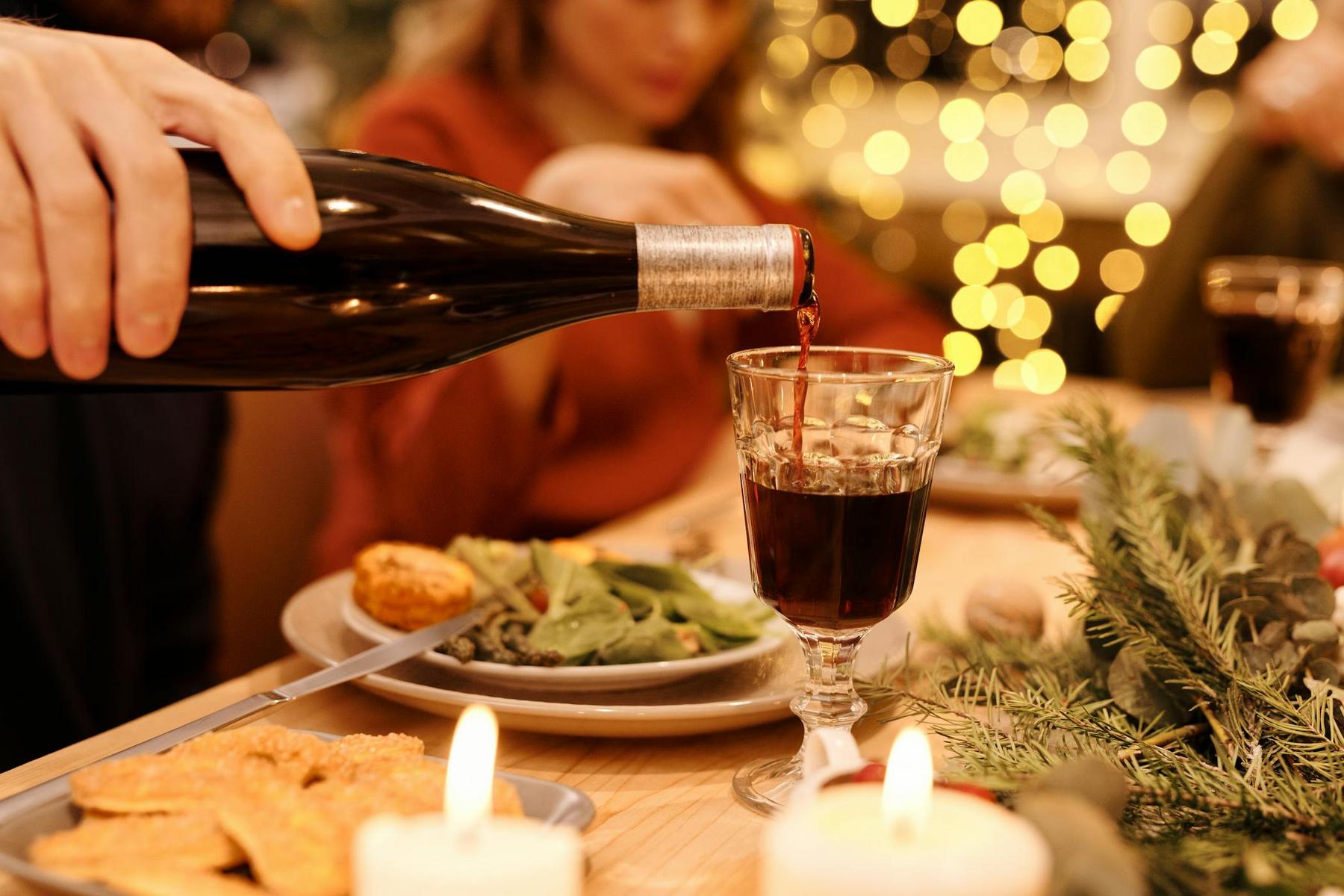 A person pours red wine into a glass at a dinner table set with food, candles, and holiday decorations.