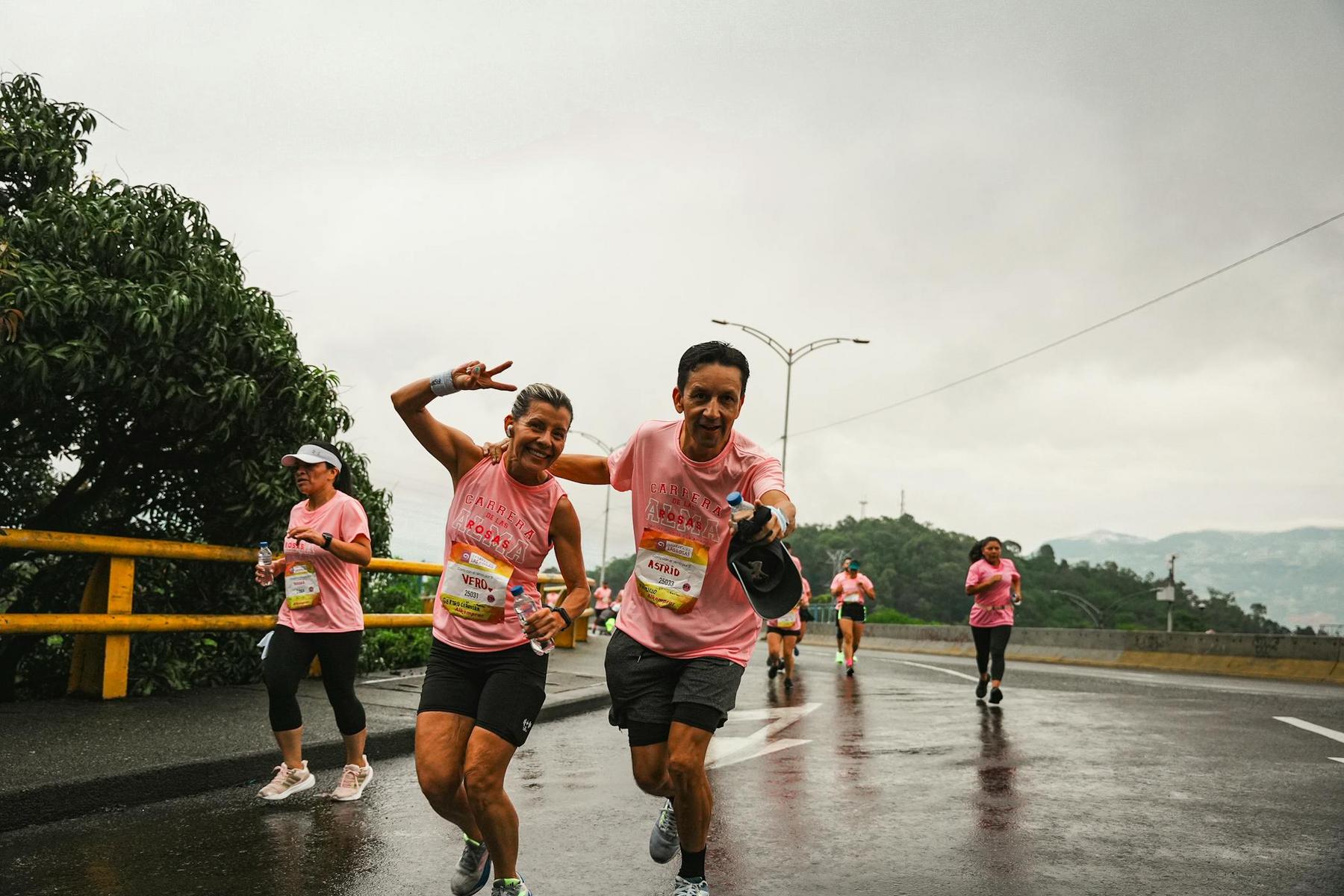 Runners in pink shirts participate in a road race on a wet, overcast day. Two people at the front smile and gesture towards the camera.