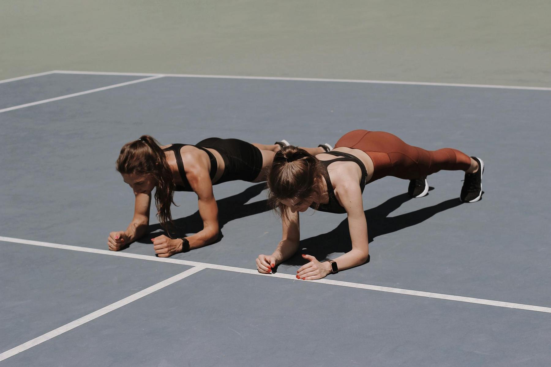 Two people wearing athletic clothing hold plank positions on a blue sports court under natural light.