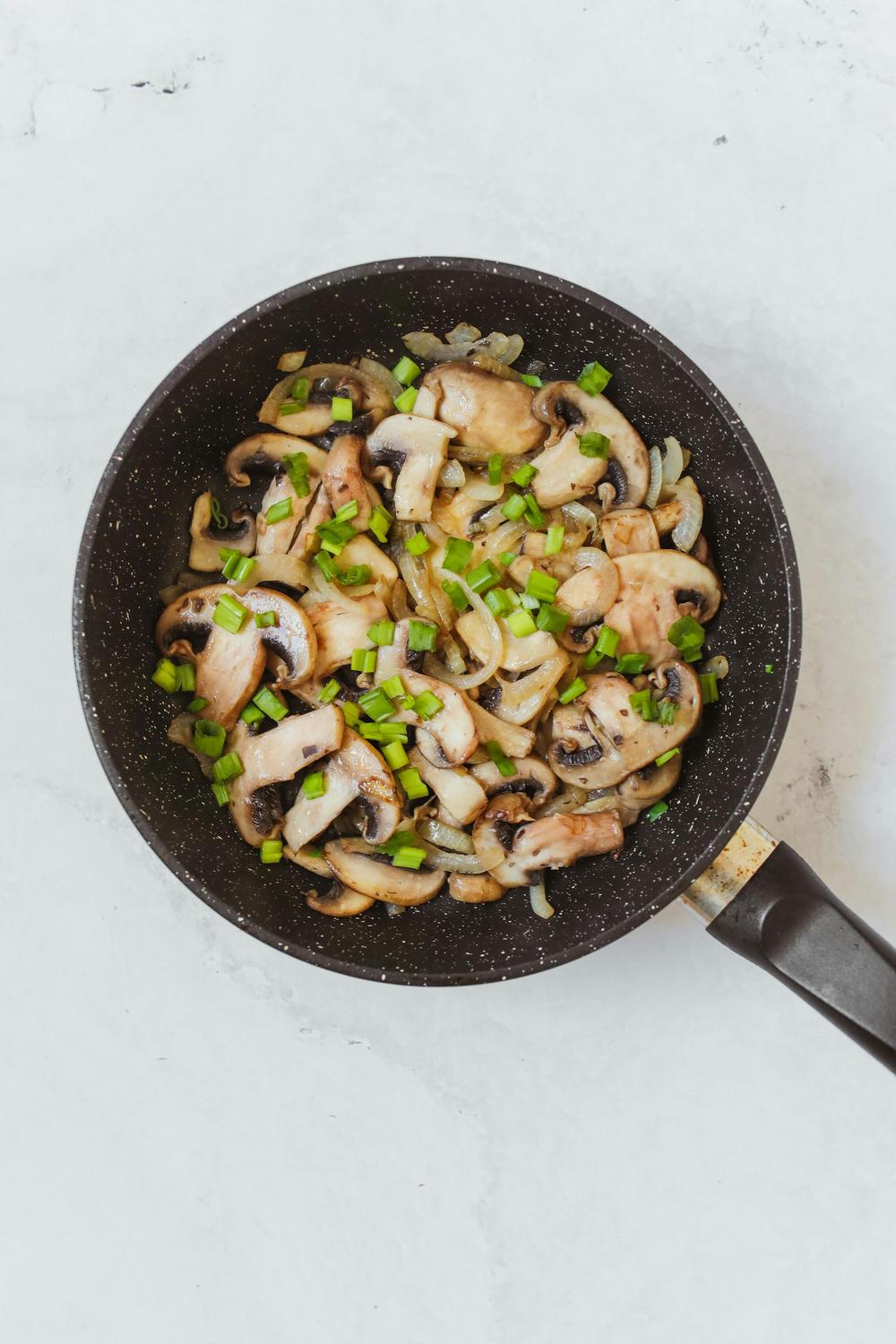 A black frying pan contains sliced mushrooms, chopped green onions, and sautéed onions on a light-colored surface.
